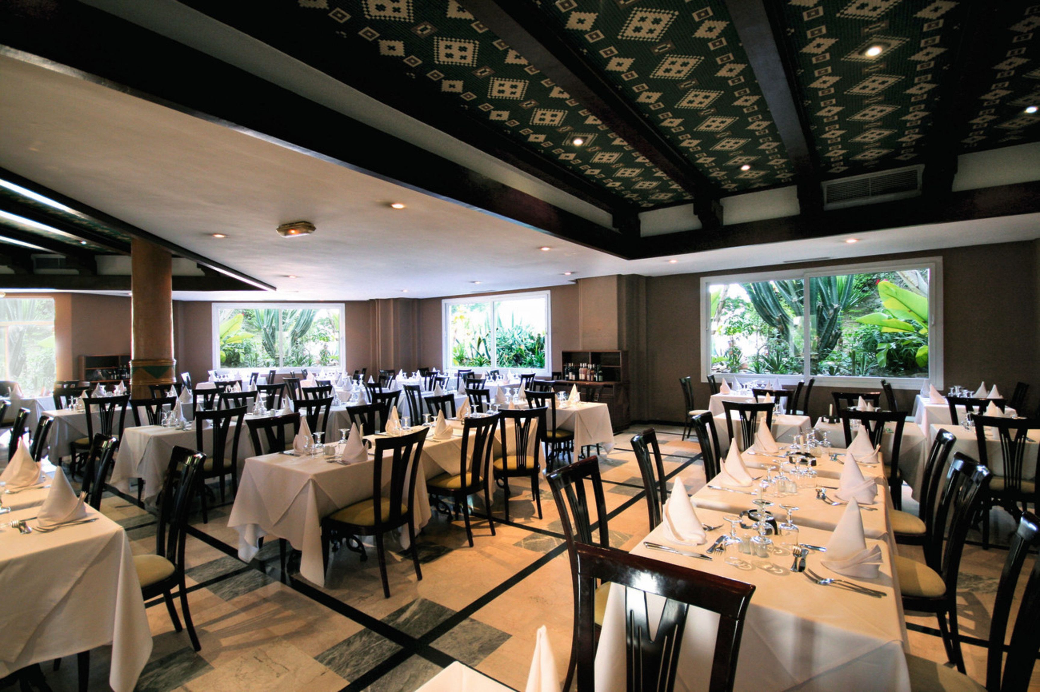 A restaurant with multiple tables set for dining, large windows with a view of greenery, and a patterned ceiling.