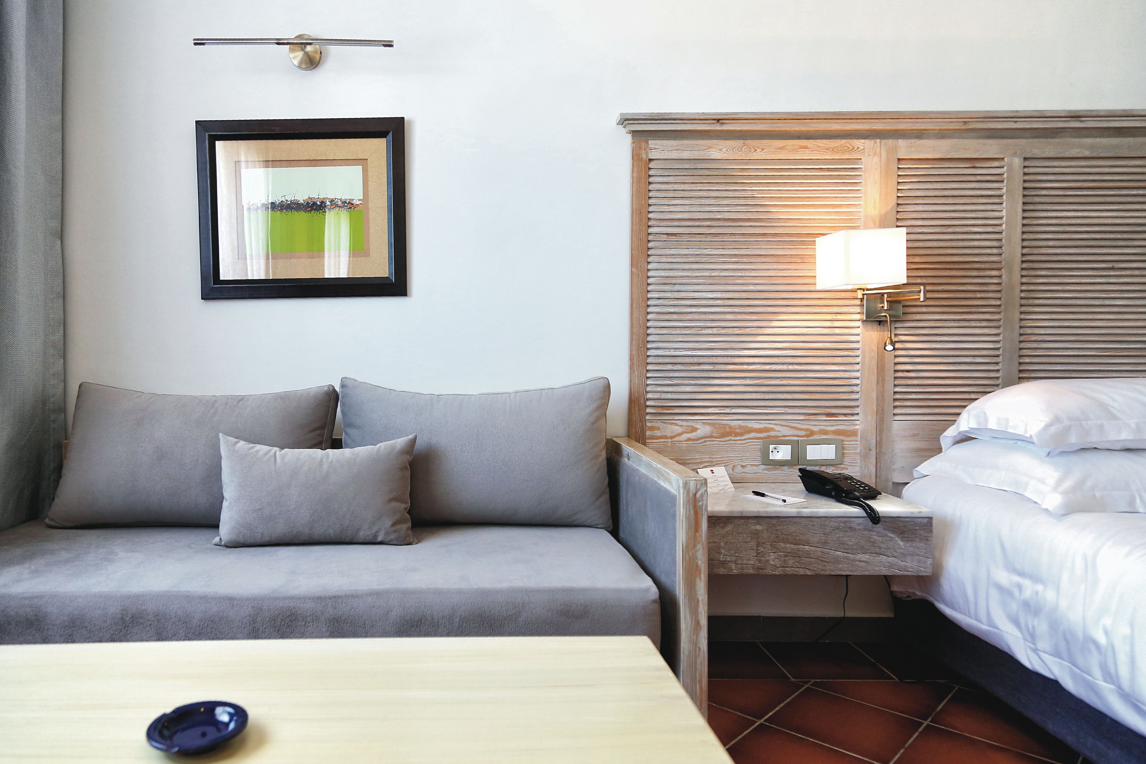 Habitación de hotel con un sofá gris, una mesita de noche con lámpara, un cuadro enmarcado en la pared y una cama con ropa blanca.