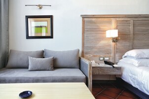 Habitación de hotel con un sofá gris, una mesita de noche con lámpara, un cuadro enmarcado en la pared y una cama con ropa blanca.