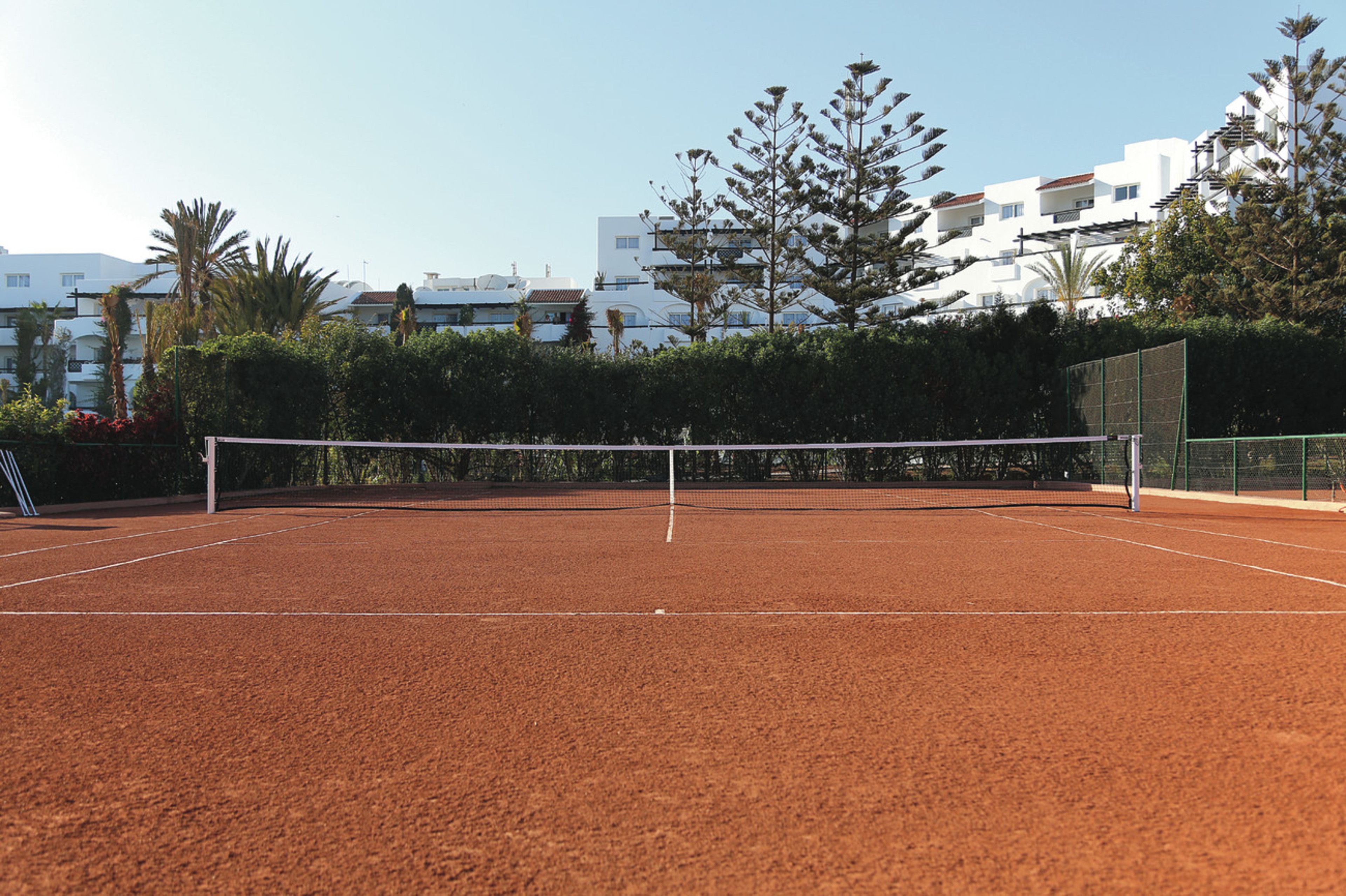 Camp de tennis amb xarxa, envoltat de vegetació i edificis al fons.