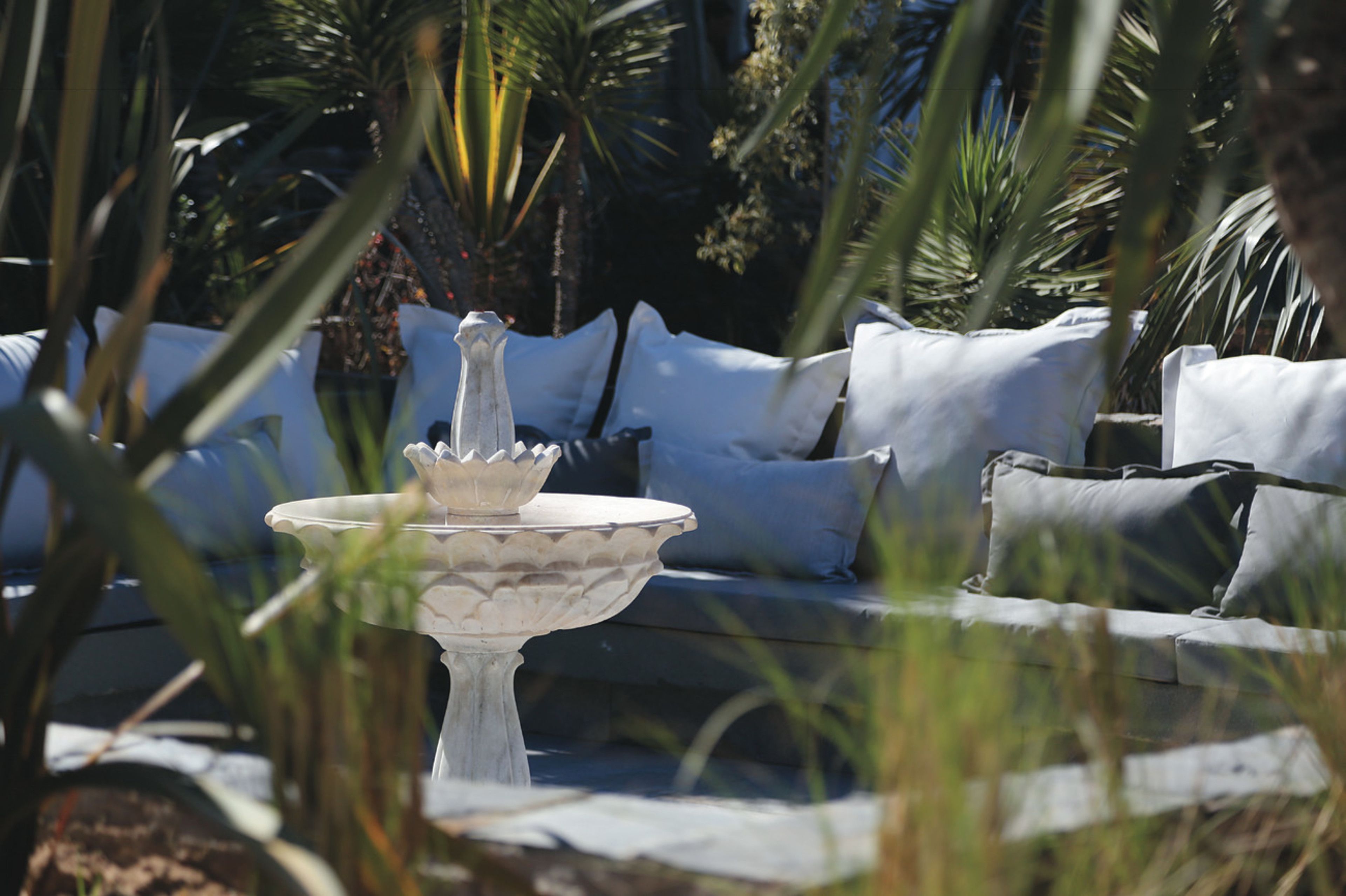 A white outdoor seating area with a stone fountain, surrounded by palm trees and greenery.