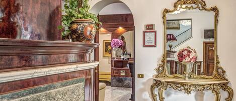A marble-floored hallway with a large ornate mirror, a potted plant, and a wooden reception desk.