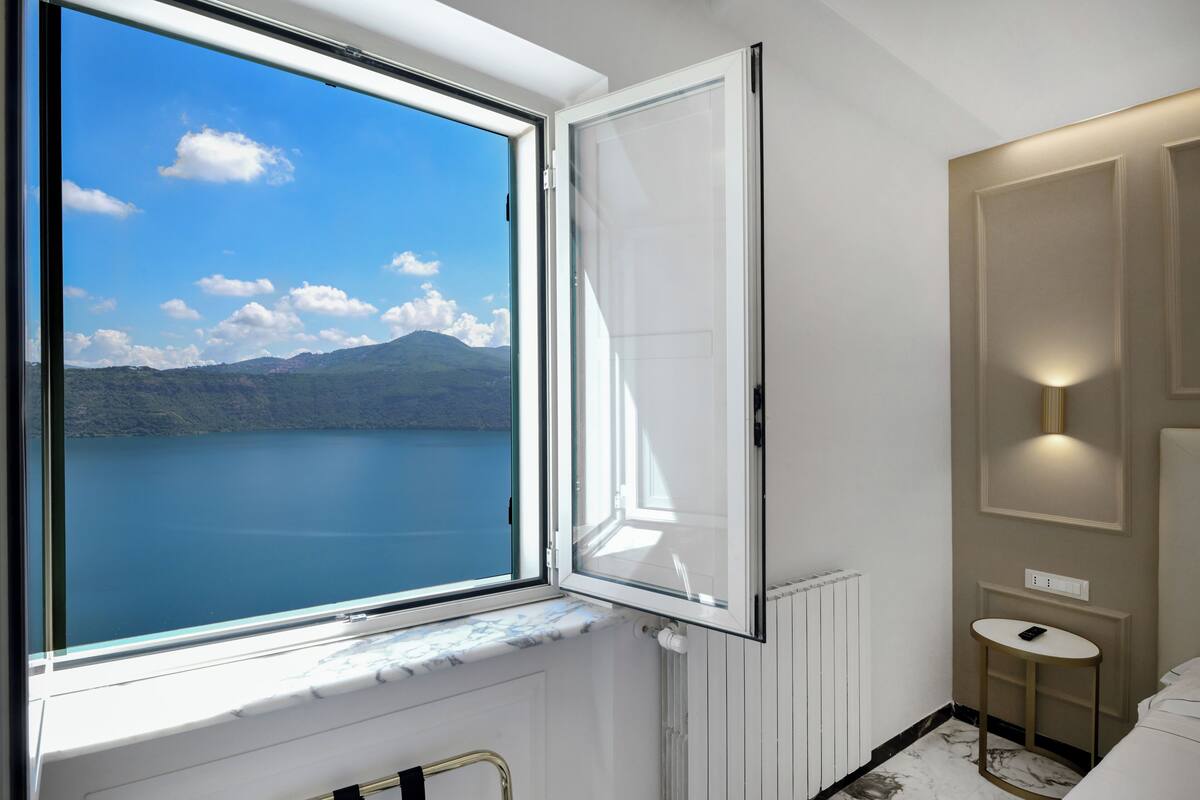 A room with a large window offering a view of a lake and mountains, a small bedside table with a lamp, and a radiator.