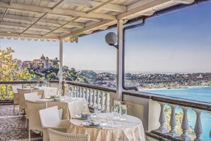 A terrace with tables set for dining, overlooking a coastal view with buildings in the distance. - Hotel Castel Vecchio (Castel Gandolfo)