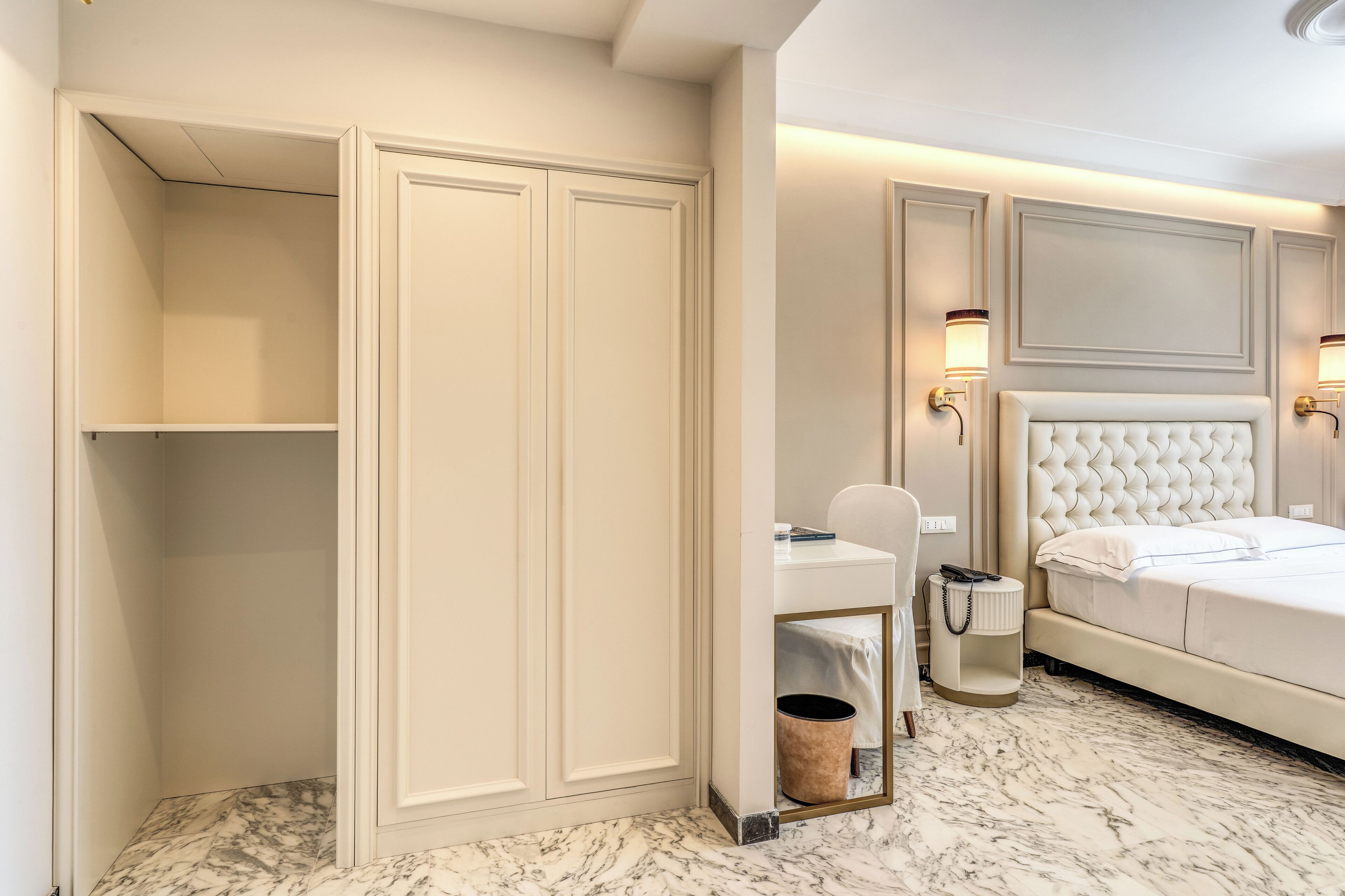 Habitación de hotel con suelo de mármol, cama blanca, mesita de noche con lámpara, escritorio con silla y armario.