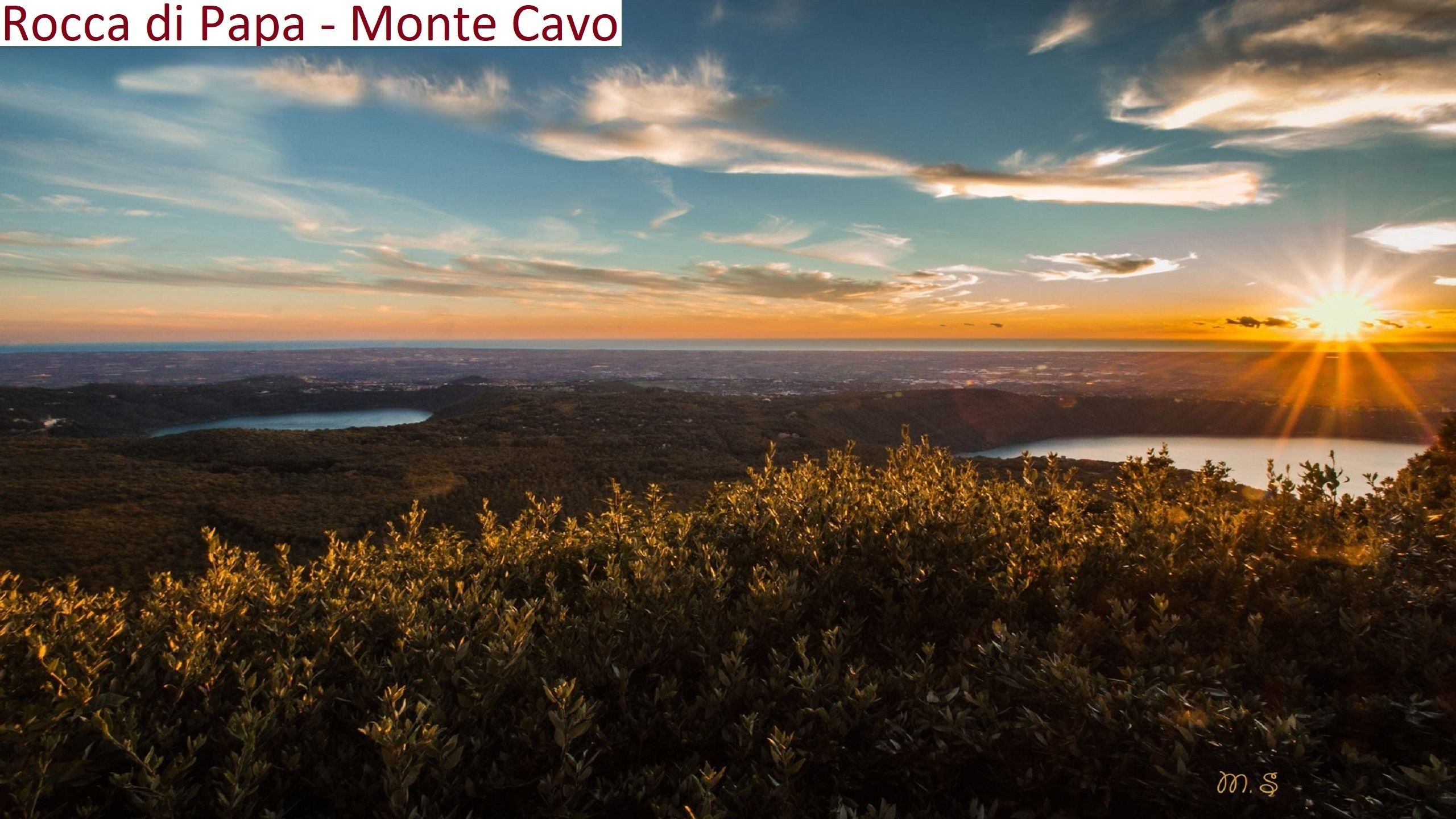 Une vue panoramique d’une région montagneuse, avec un lever ou un coucher de soleil, de la végétation au premier plan et une silhouette de ville en arrière-plan.
