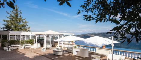 An outdoor terrace with white furniture, umbrellas, and a view of a lake.