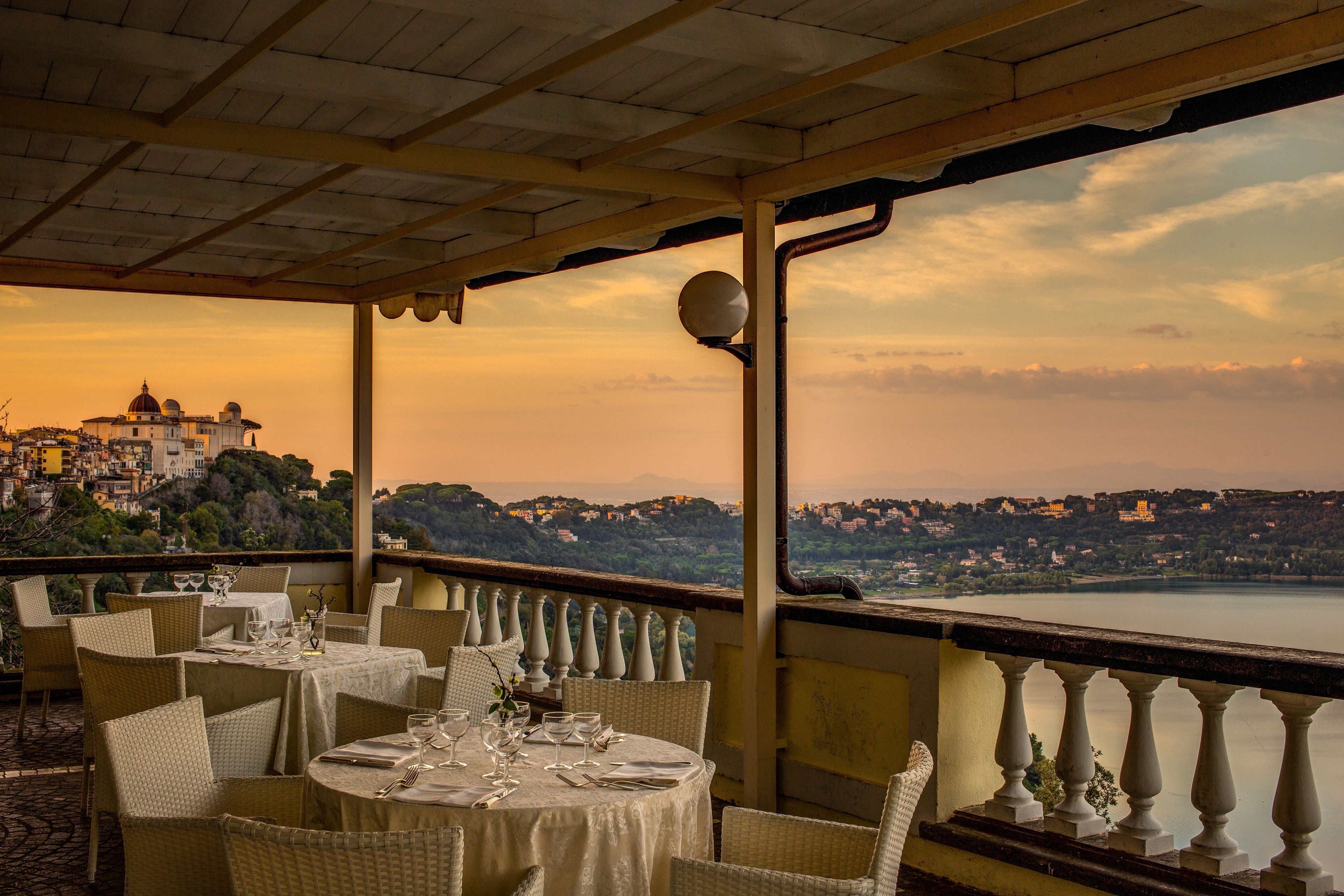 A restaurant with round tables set for dining, overlooking a scenic view of a lake and a hilltop town.