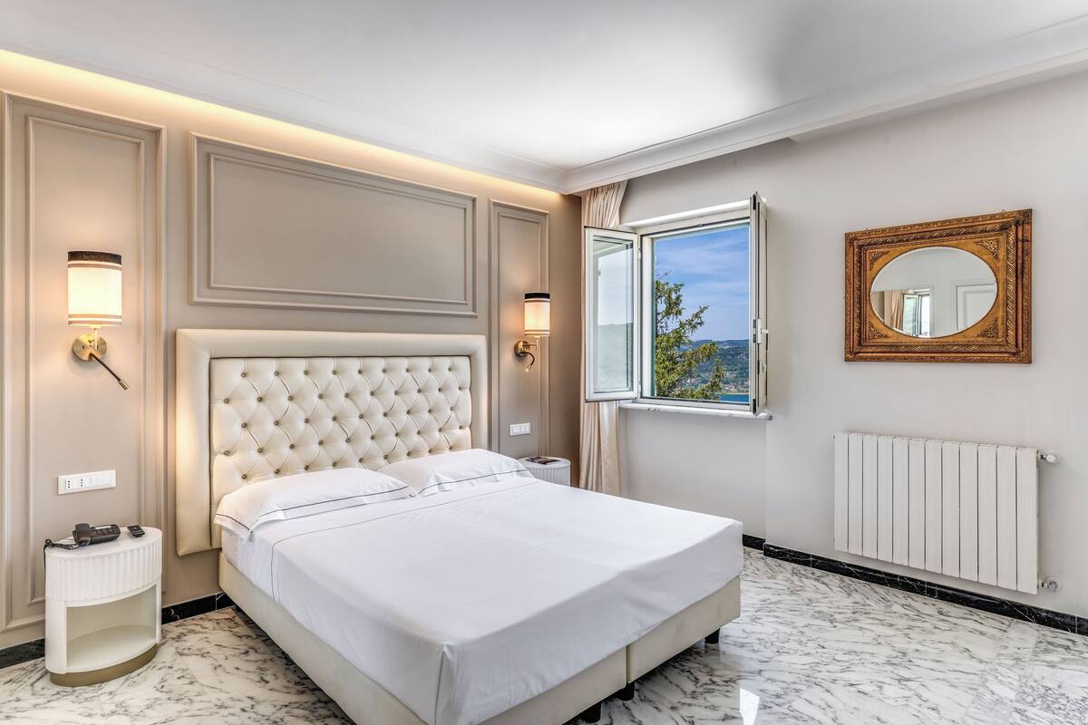 A hotel room with a large bed, a nightstand, a wall-mounted mirror, and a window with a view.
