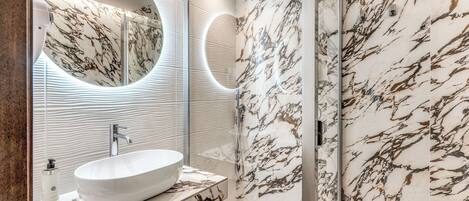 A modern bathroom with a large oval sink, a mirror, and a shower with marble walls.