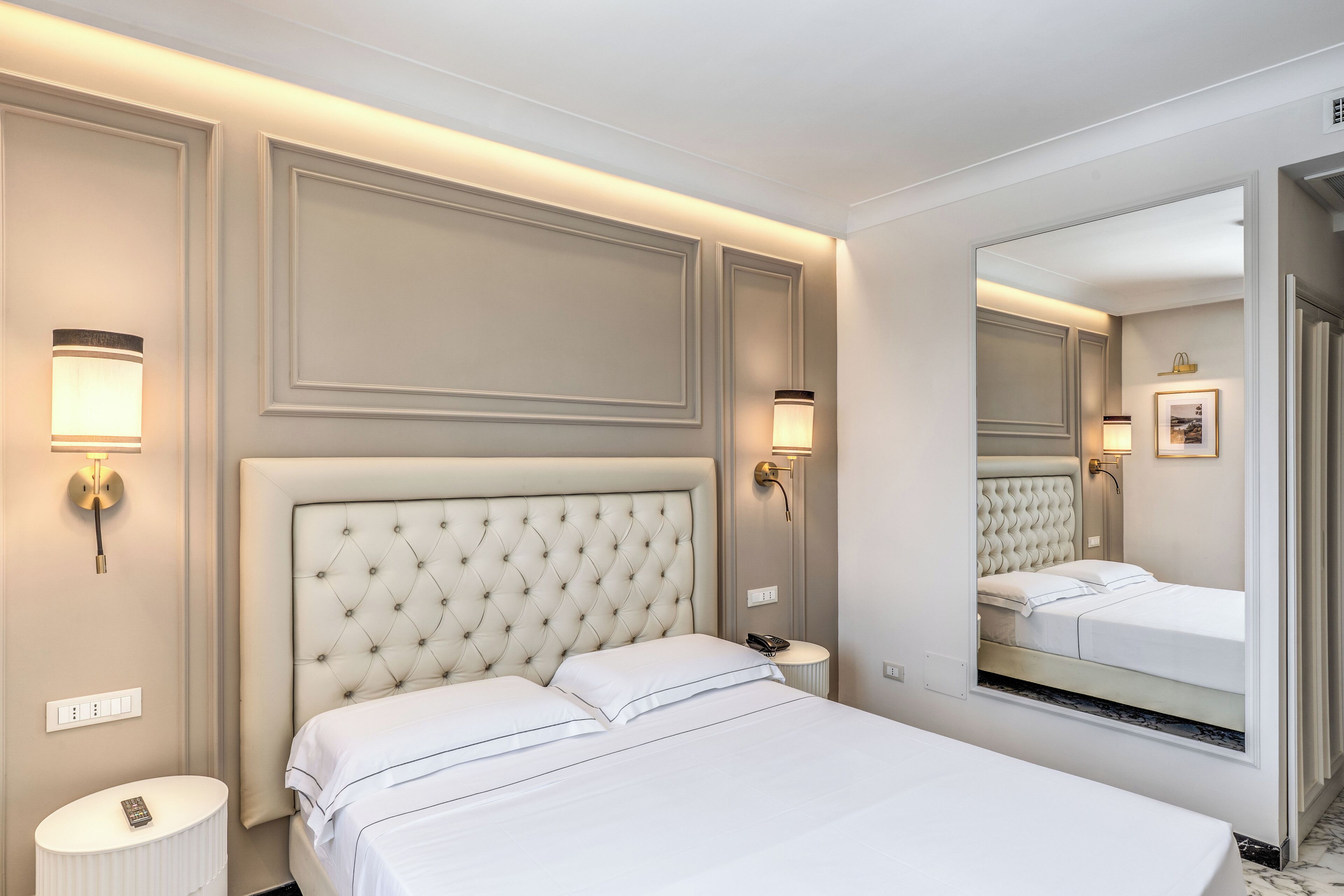 A hotel room with a large bed, a bedside table, a nightstand, a mirror, and wall-mounted lighting.