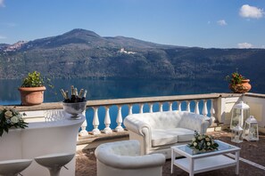 A terrace with white furniture, a view of a lake, and mountains in the background. - Hotel Castel Vecchio (Castel Gandolfo)