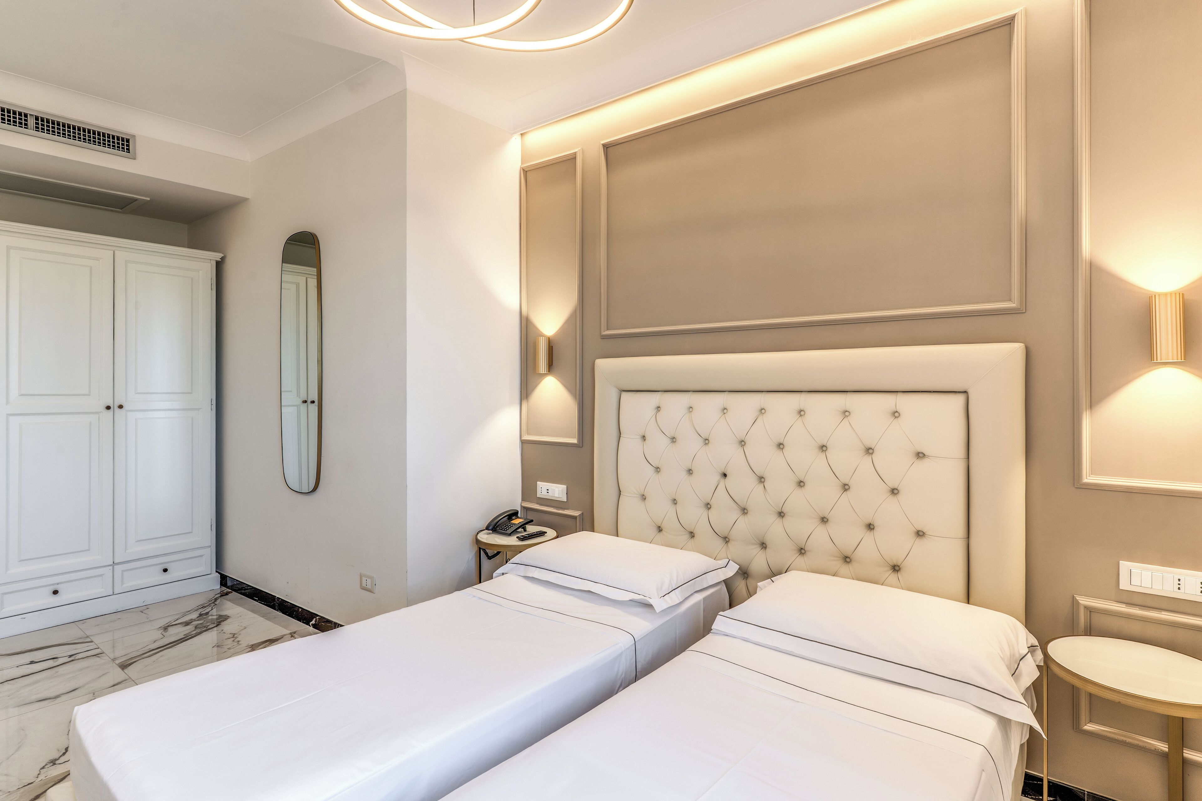A hotel room with two single beds, a wardrobe, a mirror, a bedside table with a lamp, and a wall-mounted light fixture.