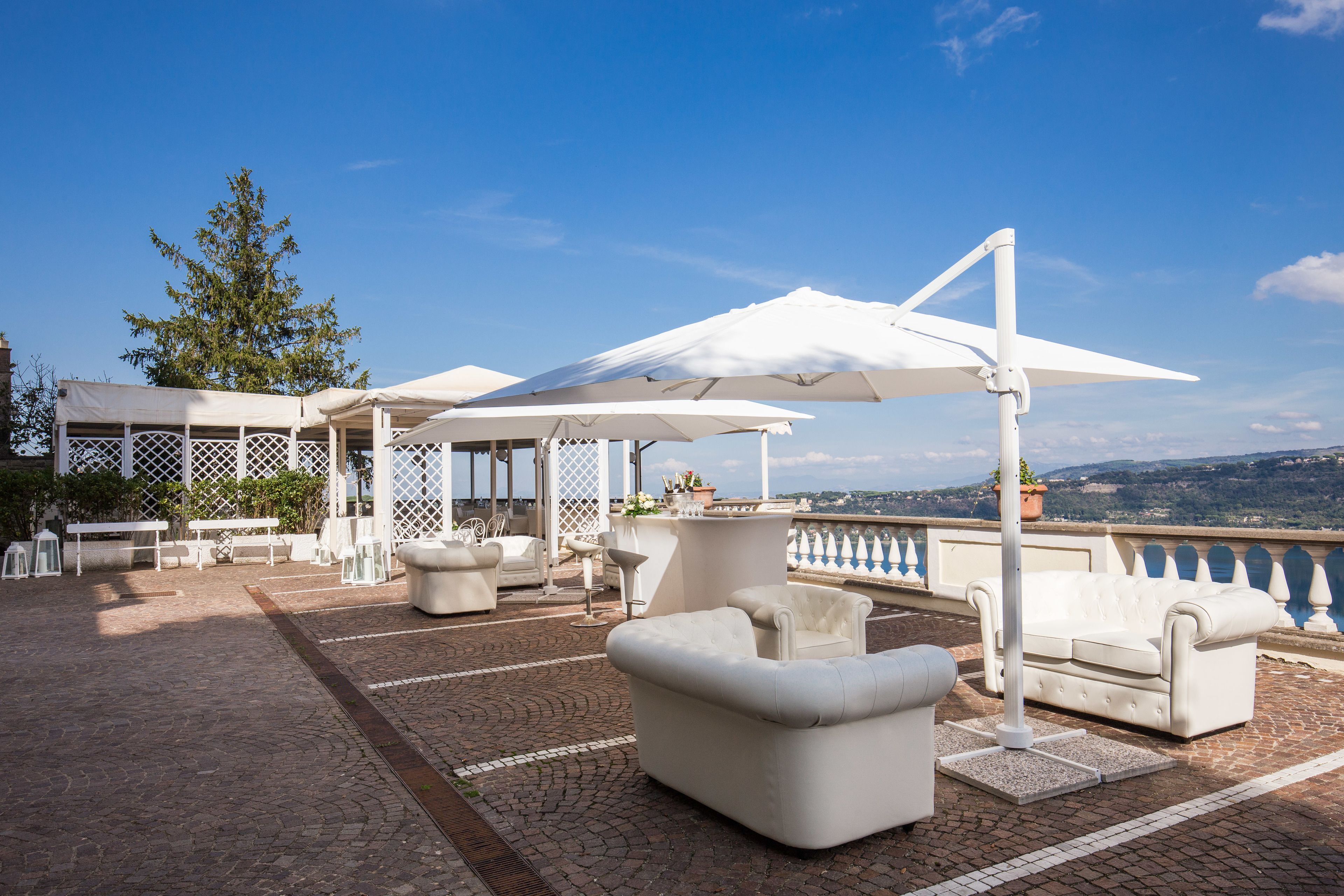 A rooftop terrace with outdoor seating, including white sofas and umbrellas, overlooking a scenic landscape.