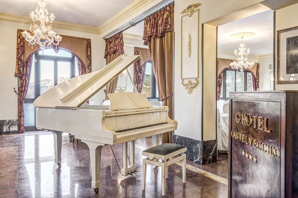 A grand piano in a well-lit room with large windows, a chandelier, and a sign reading "HOTEL CASTELVECCHIO". - Hotel Castel Vecchio (Castel Gandolfo)
