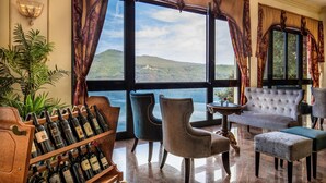 Una camera lussuosa con un'ampia finestra che offre una vista panoramica su colline e un corpo d'acqua. Si nota un portavino con bottiglie, un divano confortevole e un tavolino con un vaso.
