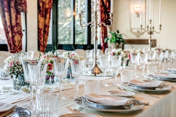 A long dining table set with glassware, plates, and napkins, with a chandelier and curtains in the background. - Hotel Castel Vecchio (Castel Gandolfo)