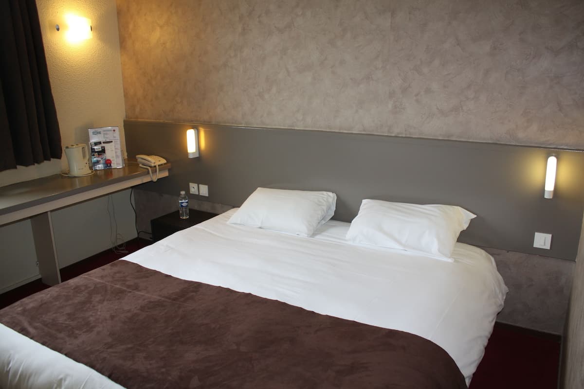 Una habitación de hotel con una cama, una mesita de noche, un teléfono y una lámpara fijada a la pared.