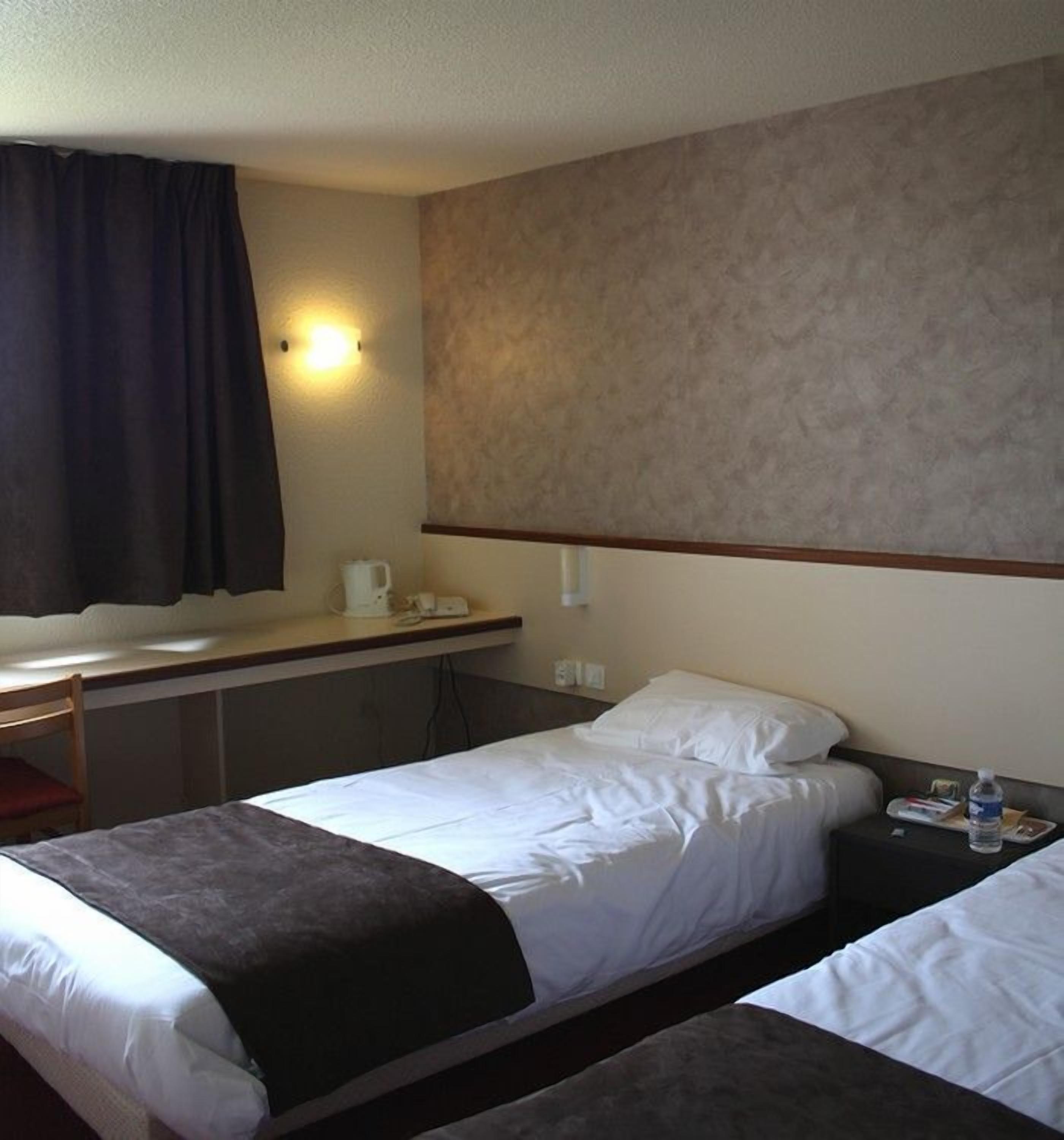 A hotel room with two single beds, a small desk with a chair, a wall-mounted light, and a window with a dark curtain.