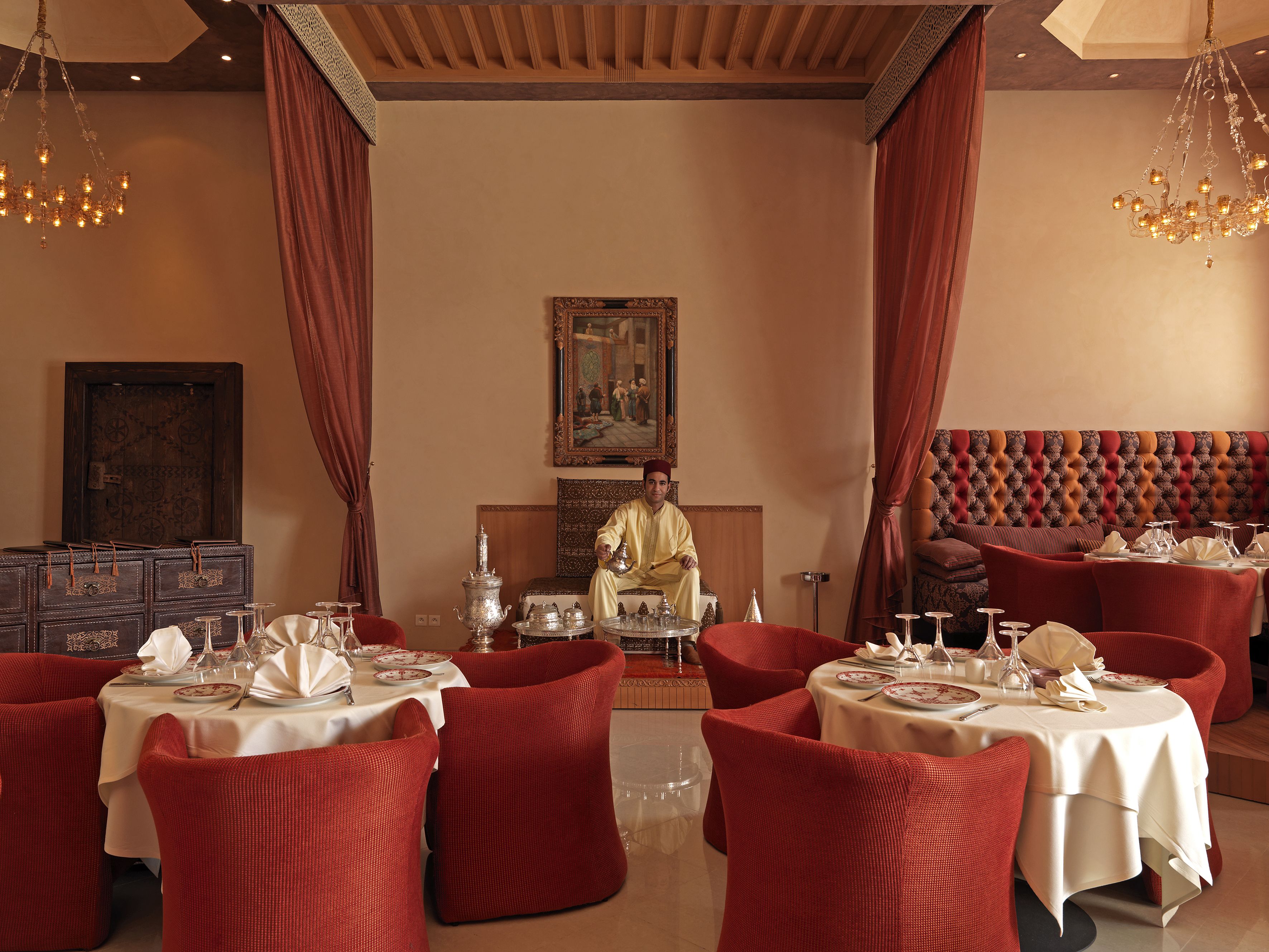 Un comedor con sillas tapizadas de rojo, mesas redondas cubiertas con manteles blancos y una araña.