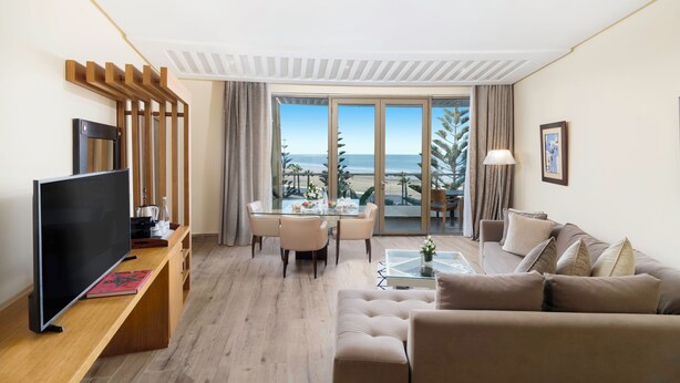 A modern hotel room with a flat-screen TV, a dining area with a table and chairs, a sofa, and a large window offering a view of the sea. - Atlas Essaouira Riad Resort (Essaouira)