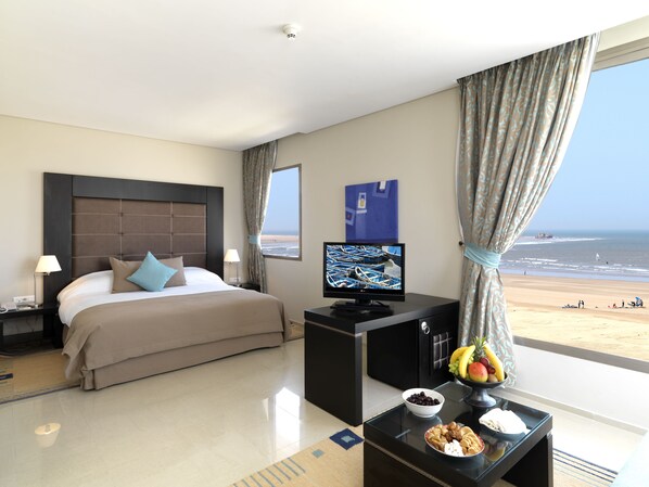 A hotel room with a bed, a desk with a computer, a view of the beach, and a fruit bowl on the desk. - Atlas Essaouira Riad Resort (Essaouira)