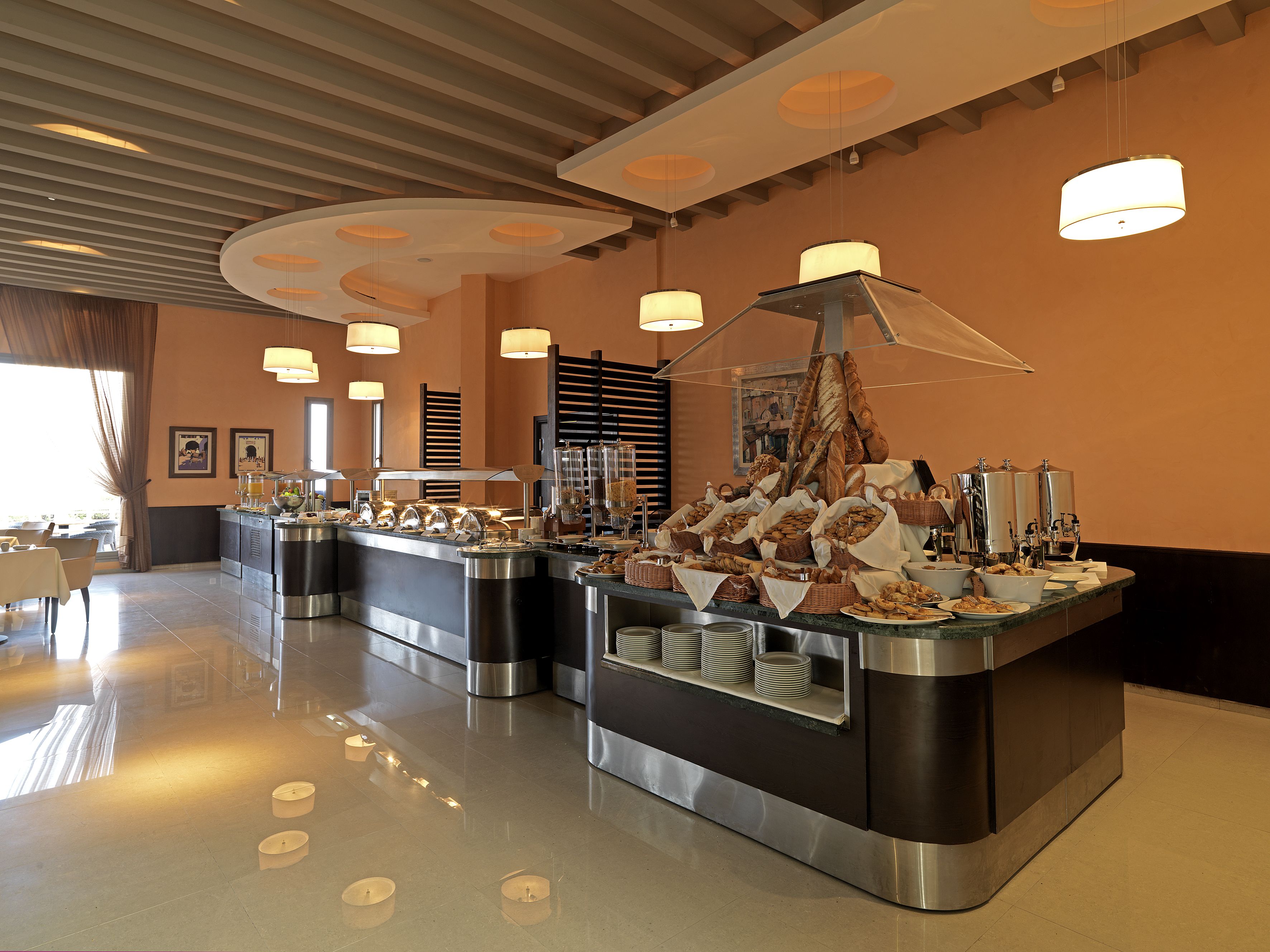 A buffet area with multiple food stations, including bread and pastries, in a modern hotel setting.