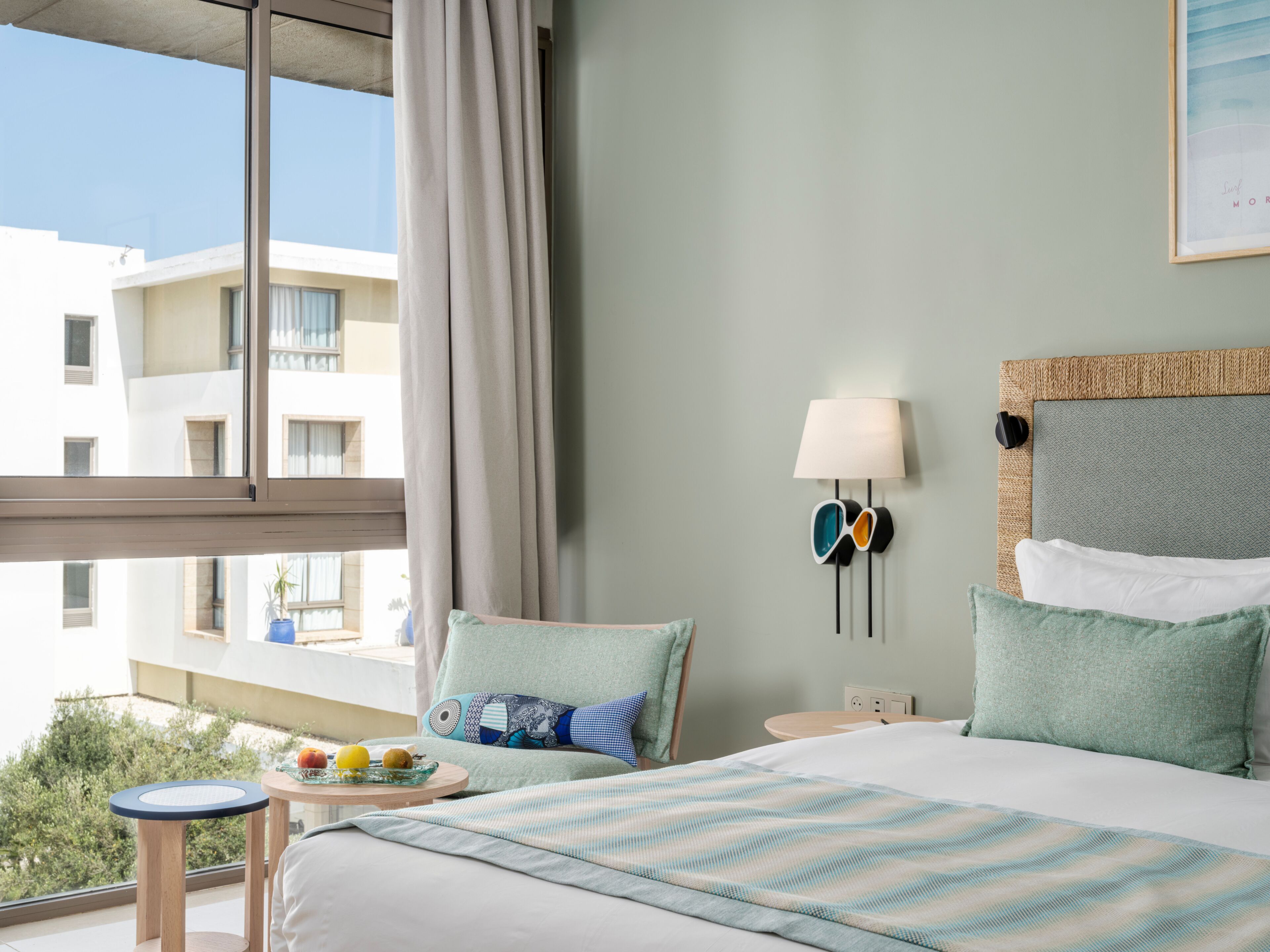 A hotel room with a bed, a small table with fruit, a lamp, and a view of buildings outside.