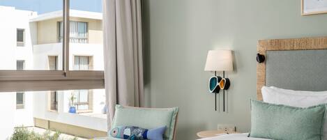 A hotel room with a bed, a small table with fruit, a lamp, and a view of buildings outside.
