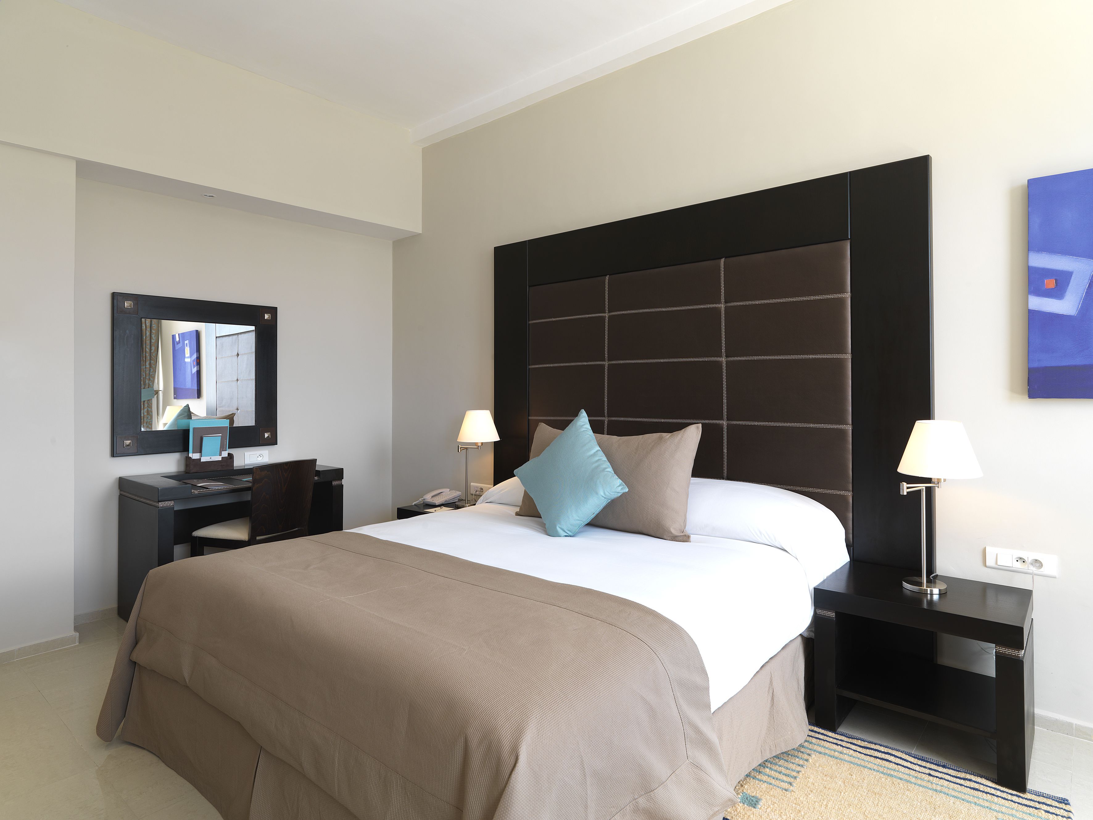 Habitación de hotel con una cama grande, un escritorio, una silla, una mesita de noche, una lámpara, un espejo y un cuadro en la pared.
