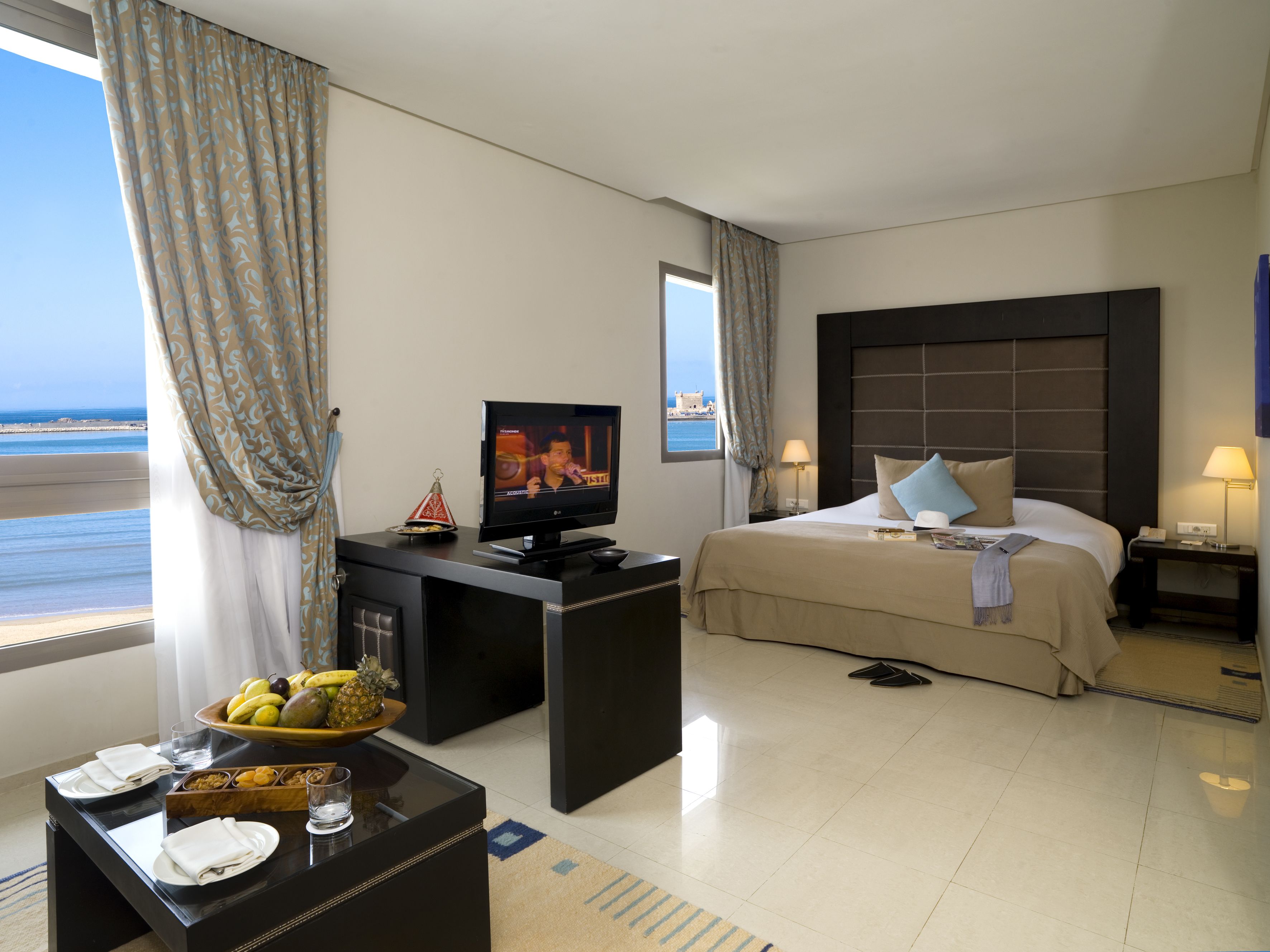 A hotel room with a bed, a TV, a desk with a fruit bowl, and a view of the sea.