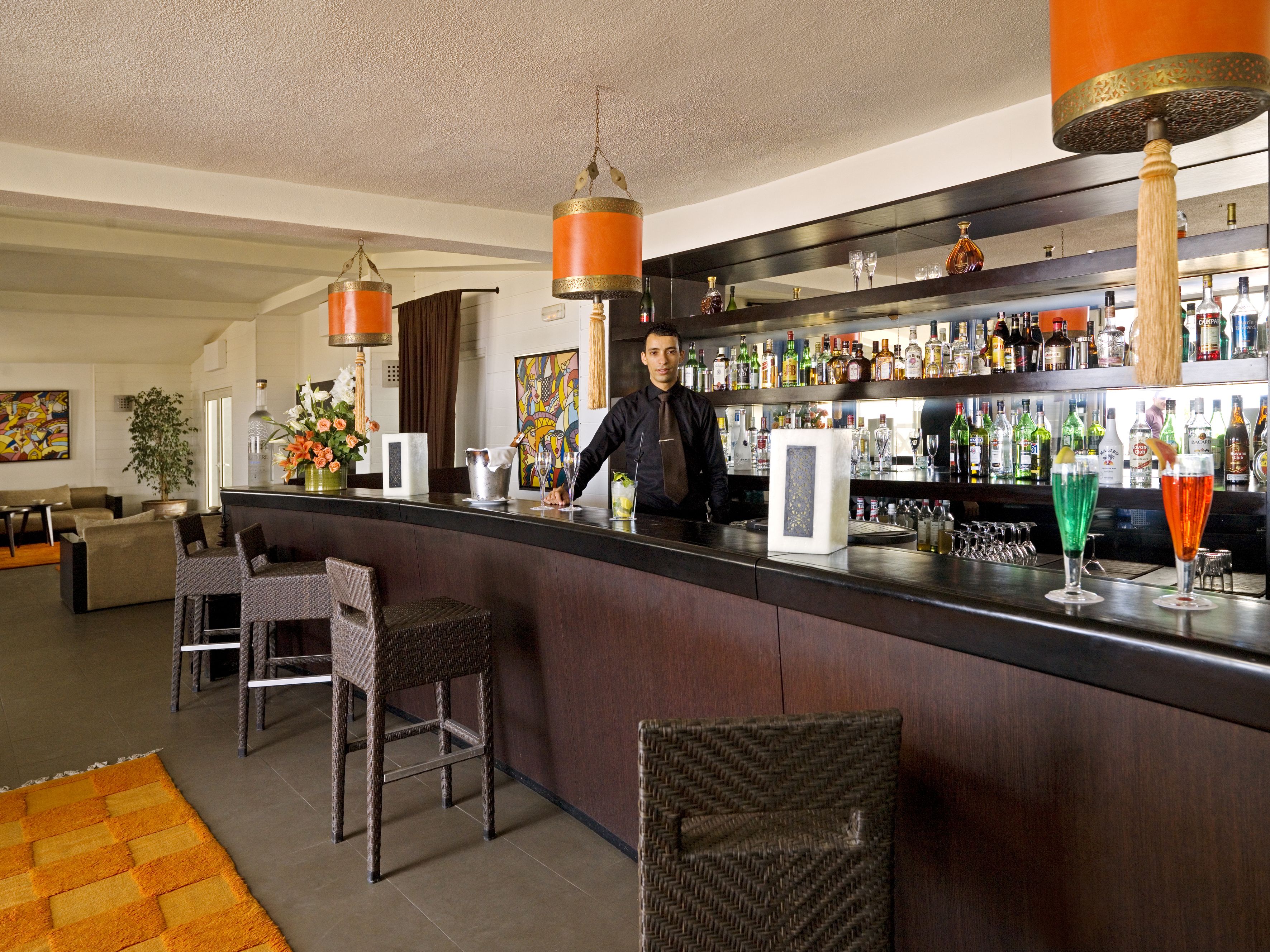 A bar area with a bartender, wicker chairs, a bar counter, and a variety of bottles and glasses.