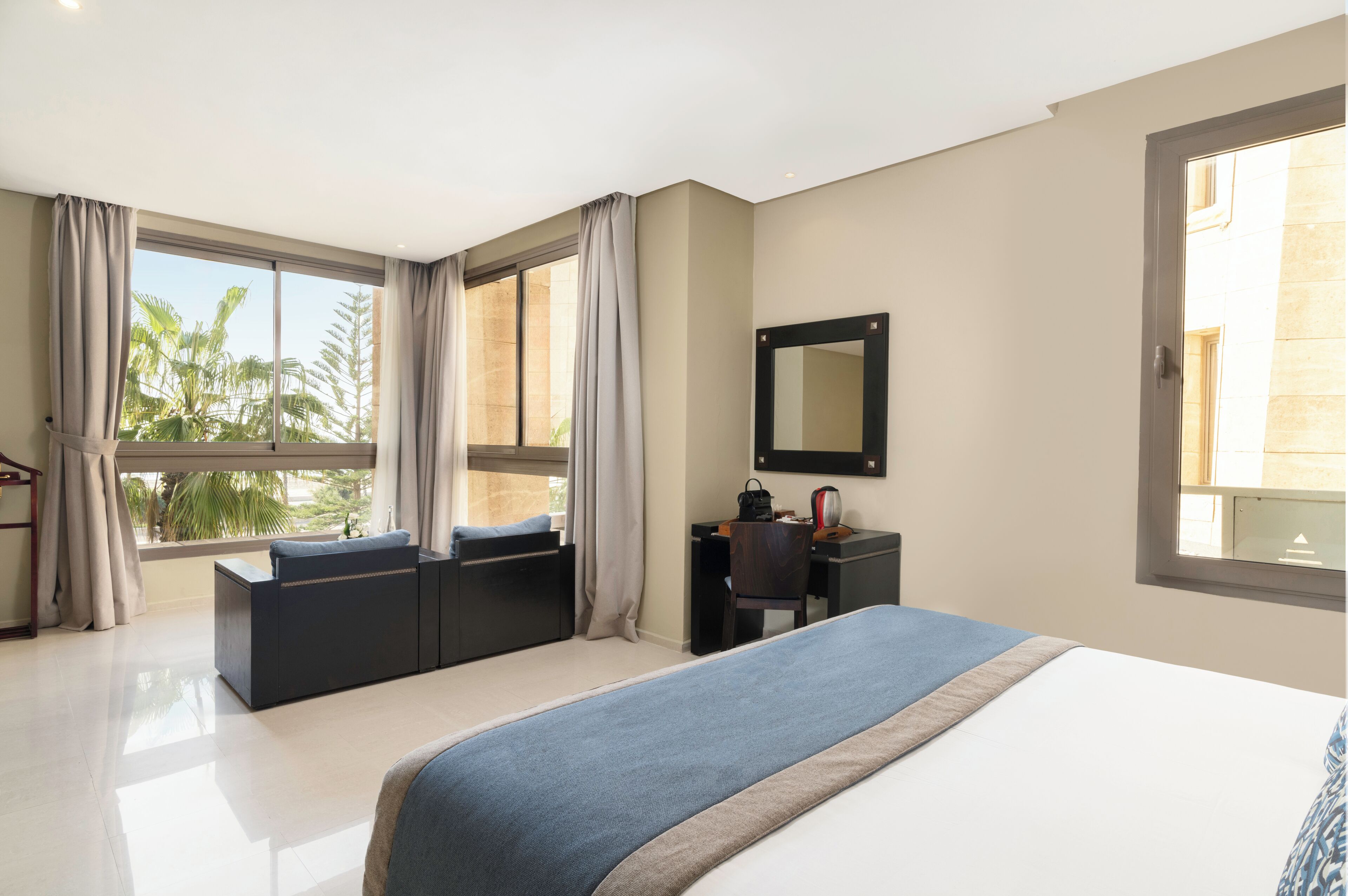 A hotel room with a large bed, a desk with a chair, a small sofa, and a large window with curtains.