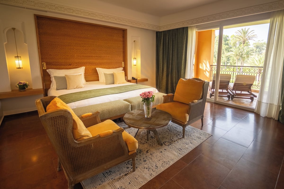 A hotel room with a large bed, two armchairs, a coffee table, and a balcony with a view of palm trees.