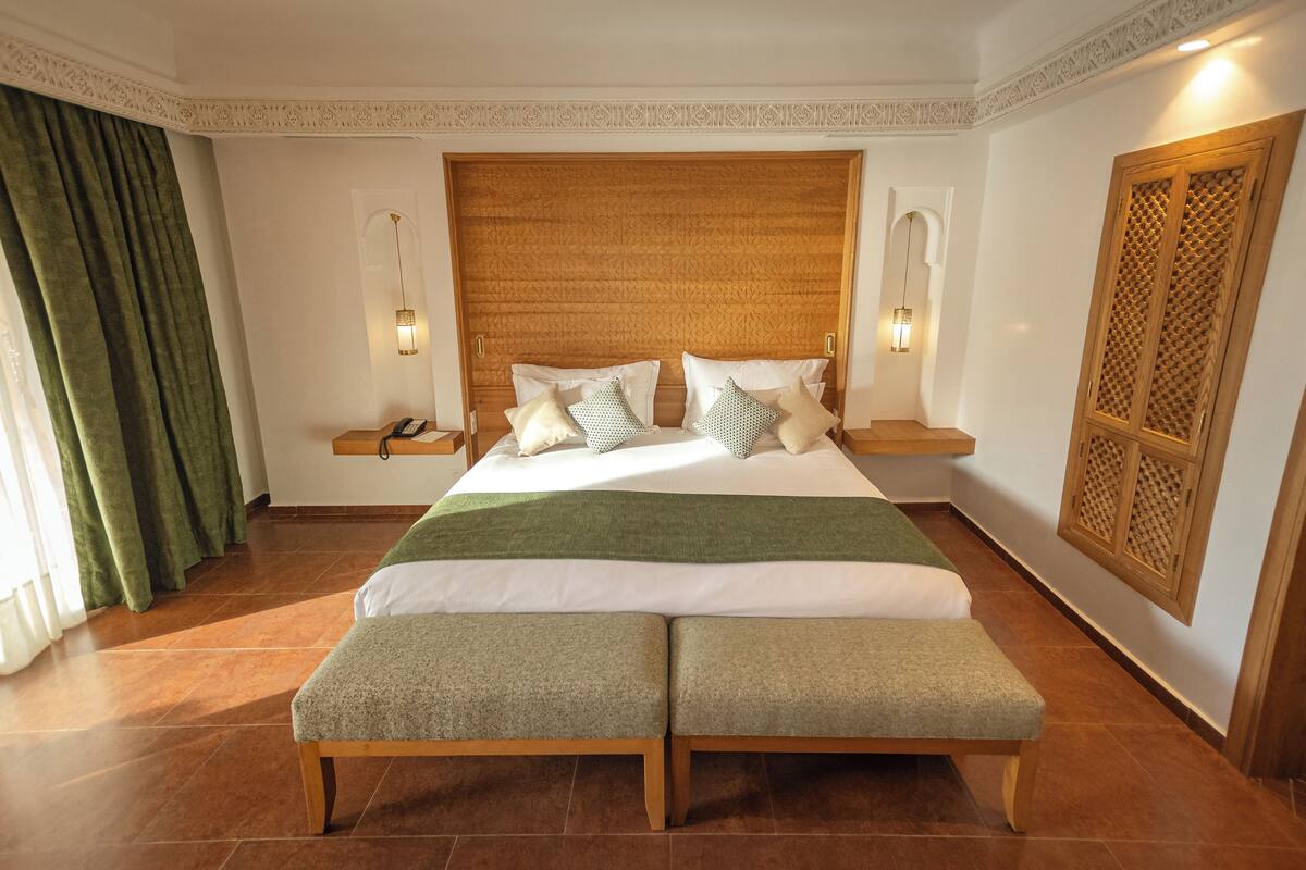 A bedroom with a large bed, two bedside tables, a bench, a decorative headboard, and a wooden panel on the wall.