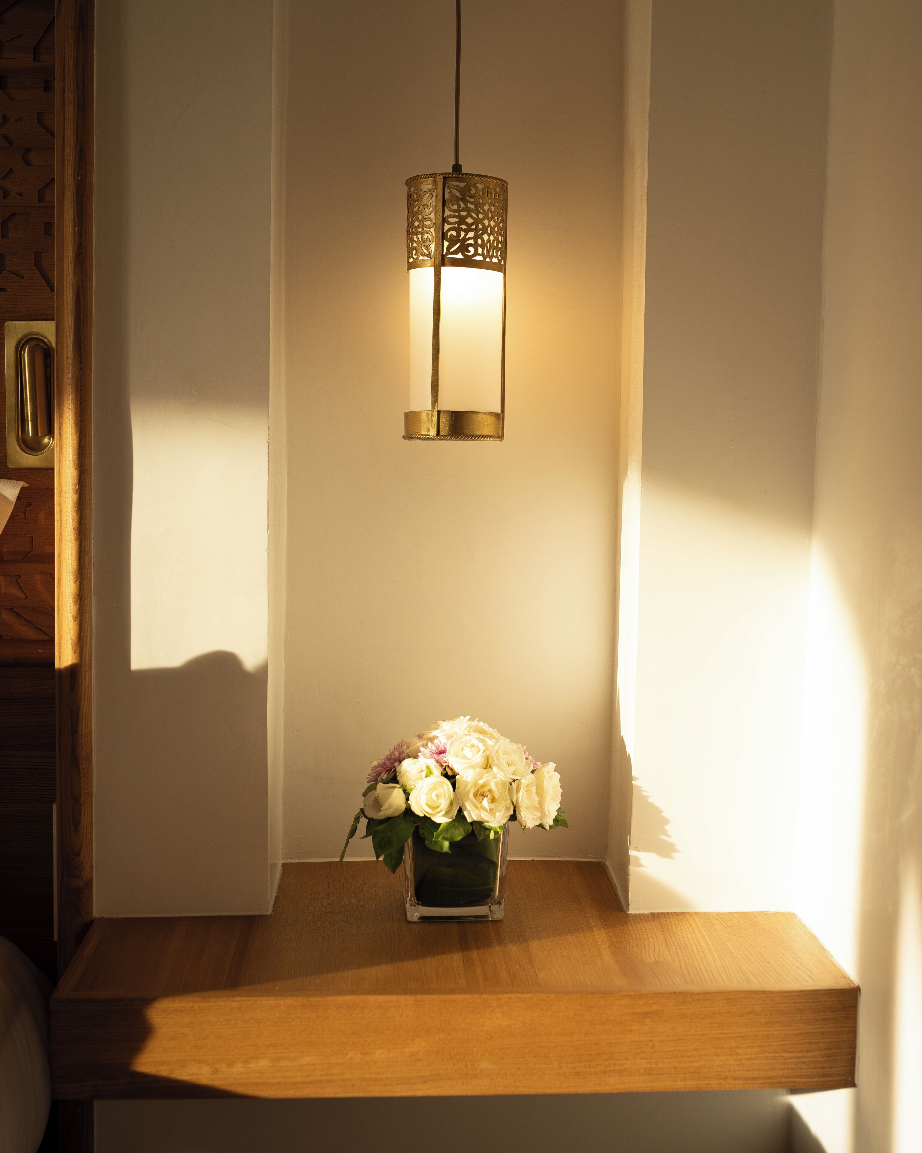 Una mesa de madera con un florero de flores blancas y una lámpara fijada a la pared.