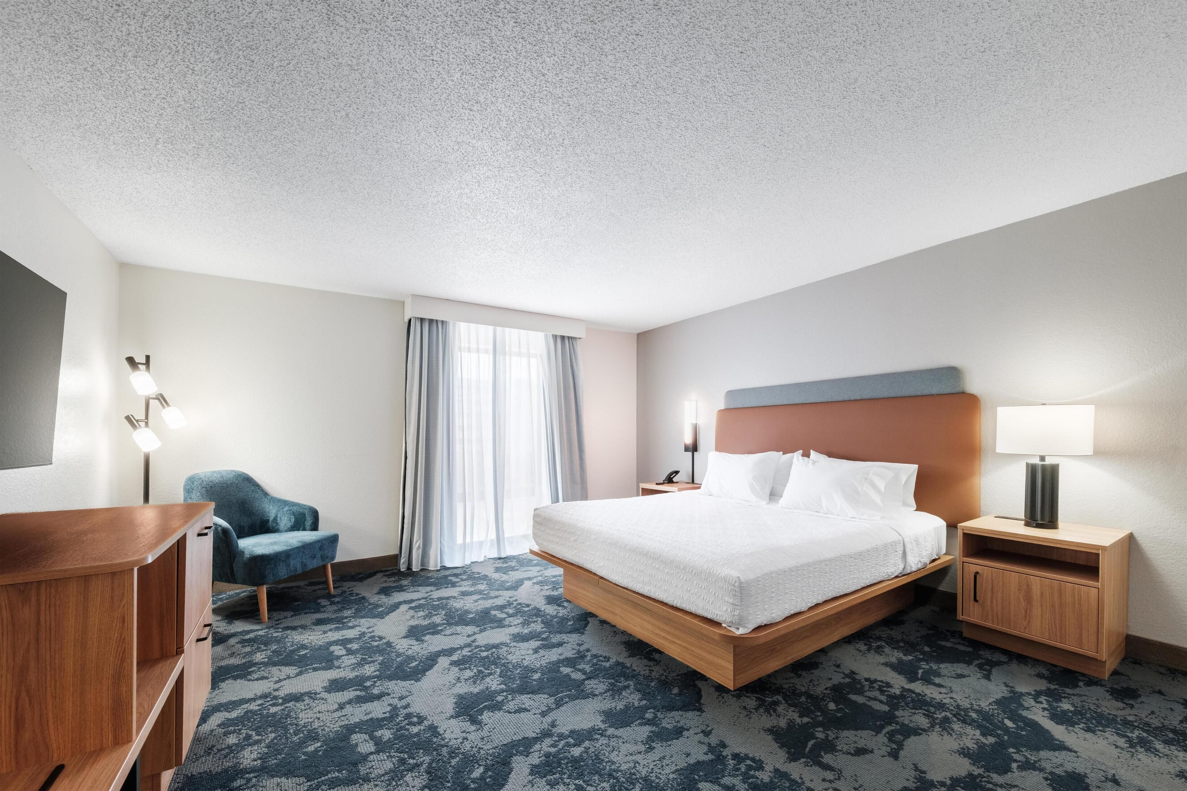 A hotel room with a bed, bedside table, lamp, chair, and a desk.