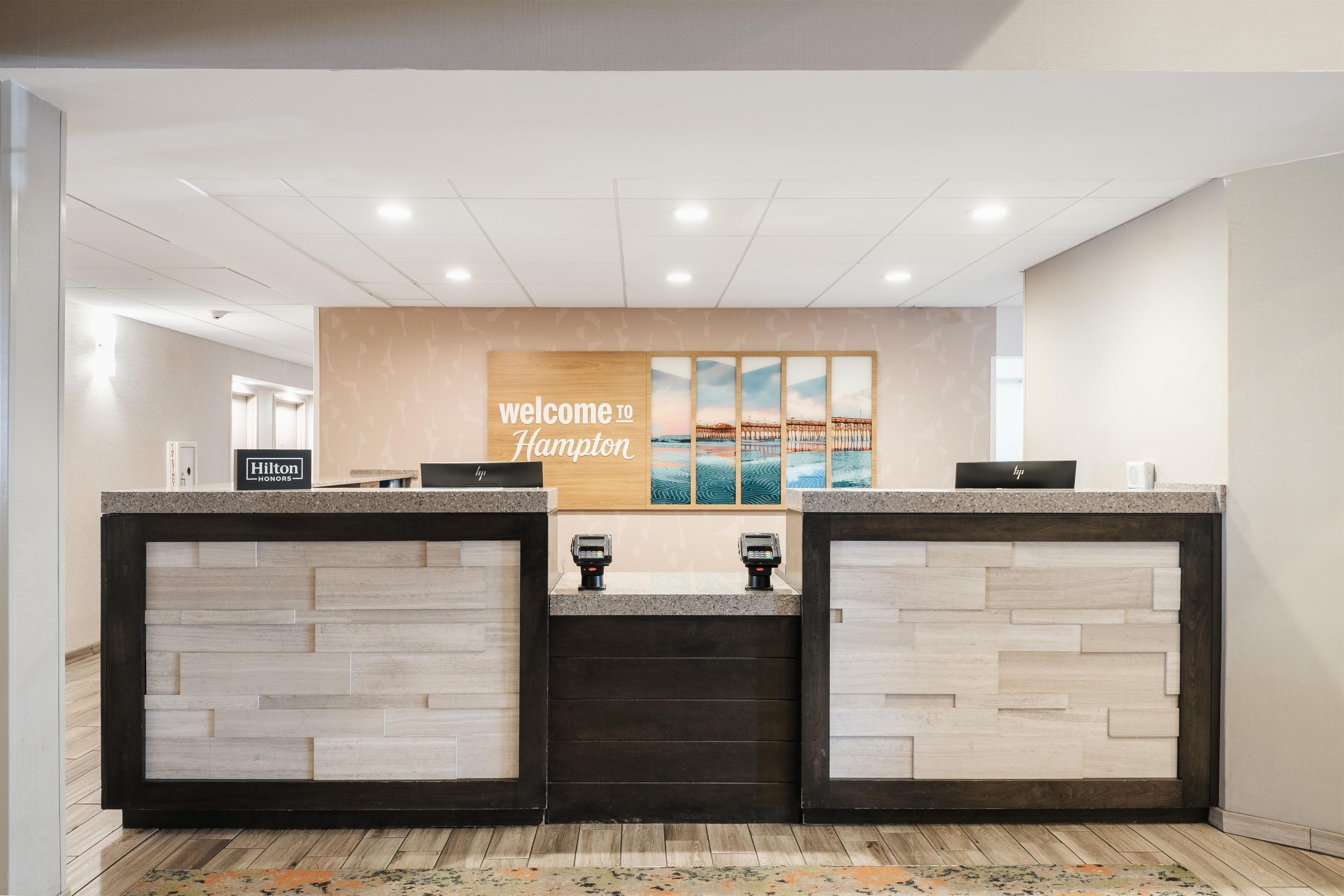 Una mesa de recepción con un cartel que dice 'Welcome to Hampton', una placa de 'Hilton Honors' y vista al océano a través de una ventana.