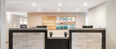 Una mesa de recepción con un cartel que dice 'Welcome to Hampton', una placa de 'Hilton Honors' y vista al océano a través de una ventana.