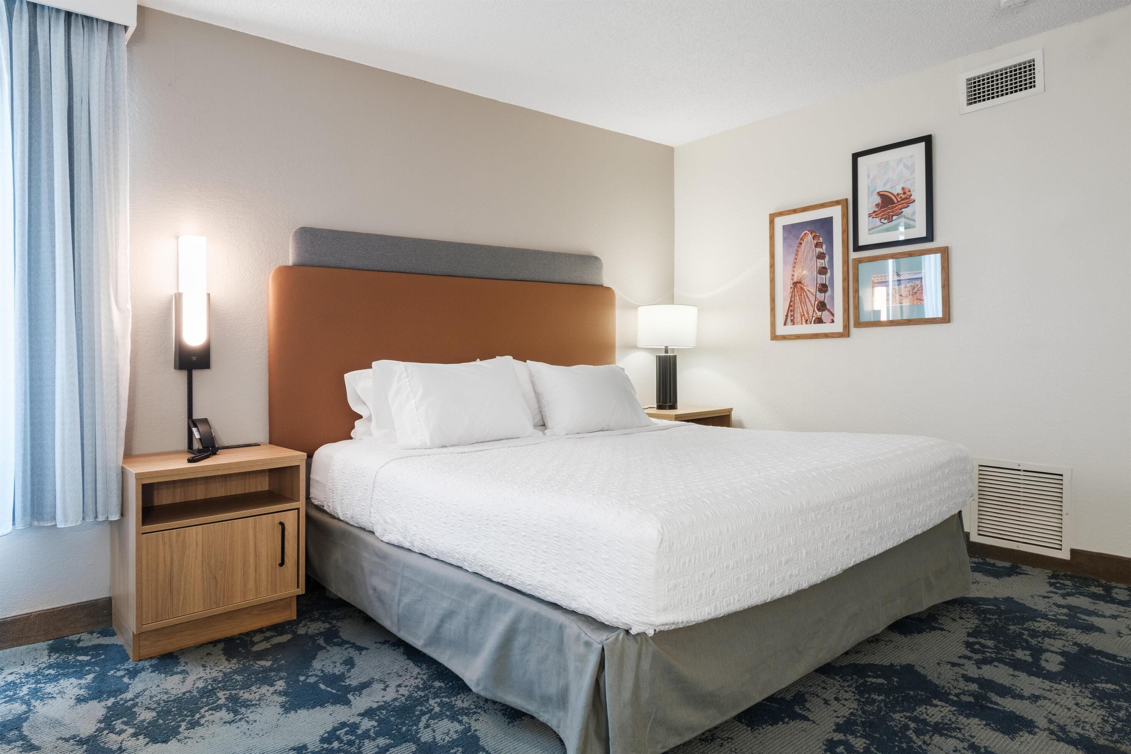A hotel room with a bed, bedside tables, a nightstand, a lamp, and framed pictures on the wall.
