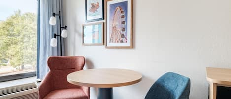 A hotel room with a round table, two chairs, a window with curtains, and framed artwork on the wall.