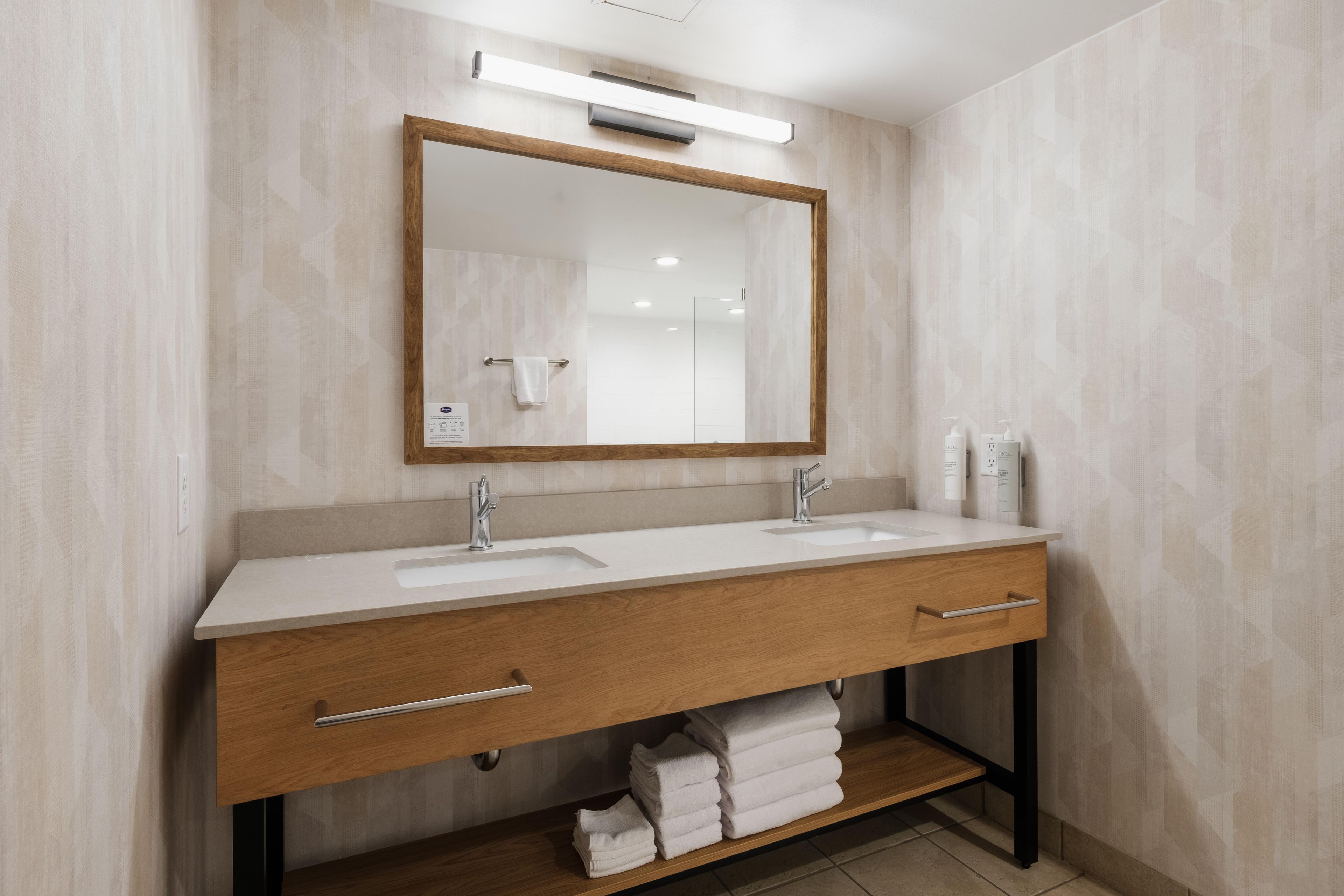 Une salle de bain moderne avec un meuble-lavabo en bois, deux vasques, un grand miroir et une étagère à serviettes.