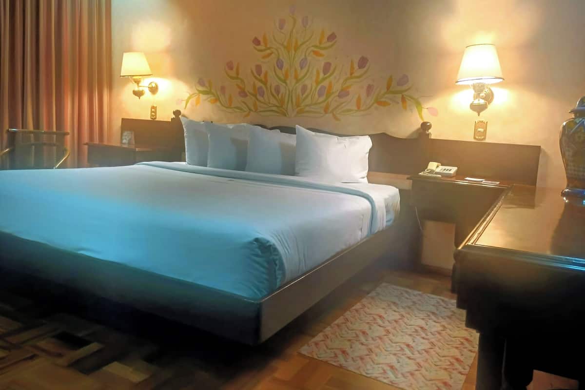 Una habitación de hotel con una cama grande, dos mesitas de noche con lámparas, una mesita con un teléfono, un elemento decorativo en la pared y suelo de madera.