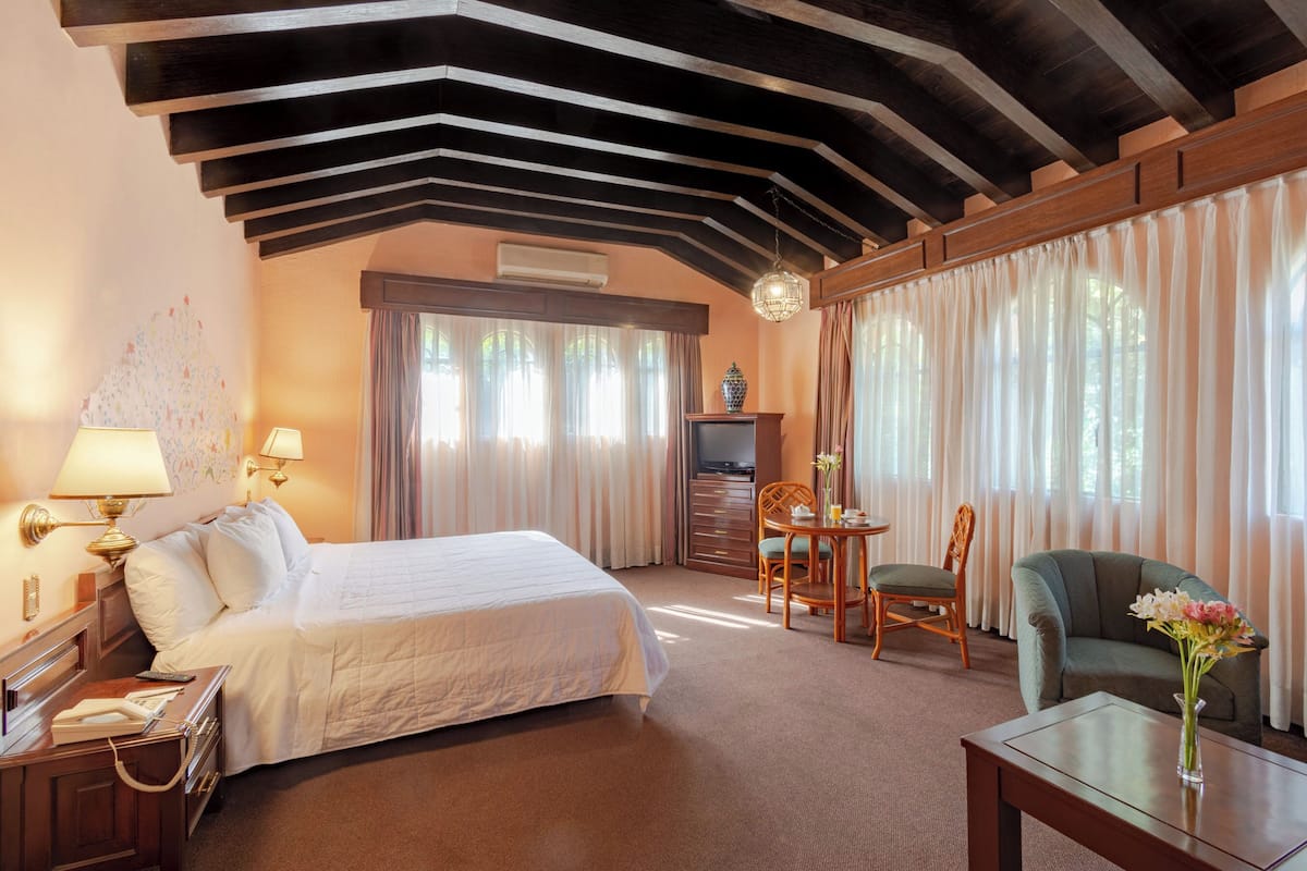 Una habitación de hotel con una cama grande, un escritorio con lámpara, una silla, un sofá y un florero con flores.