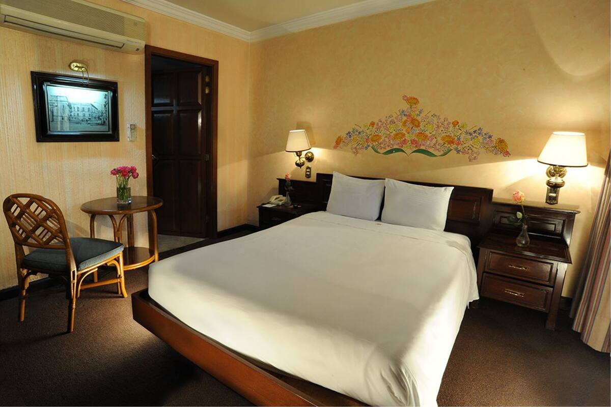 Habitación de hotel con una cama grande, dos mesitas de noche con lámparas, una silla, una pequeña mesa redonda con un jarrón de flores y un cuadro en la pared.
