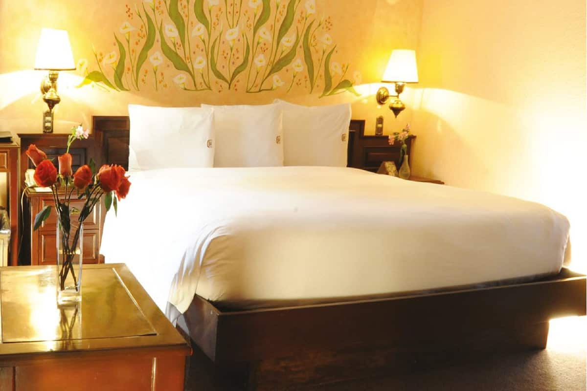 Habitación de hotel con una cama grande, dos lámparas de noche, una mesita de noche, un jarrón con flores y un mural en la pared.
