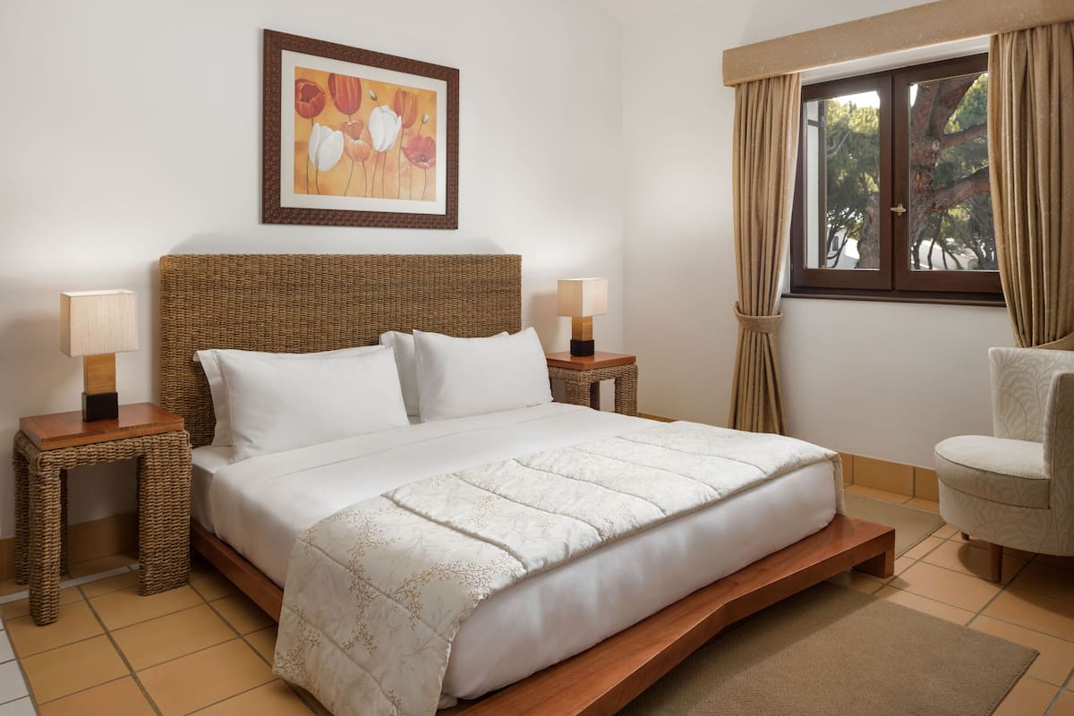 A neatly arranged bedroom with a large bed, two bedside tables with lamps, a chair, and a window with curtains.