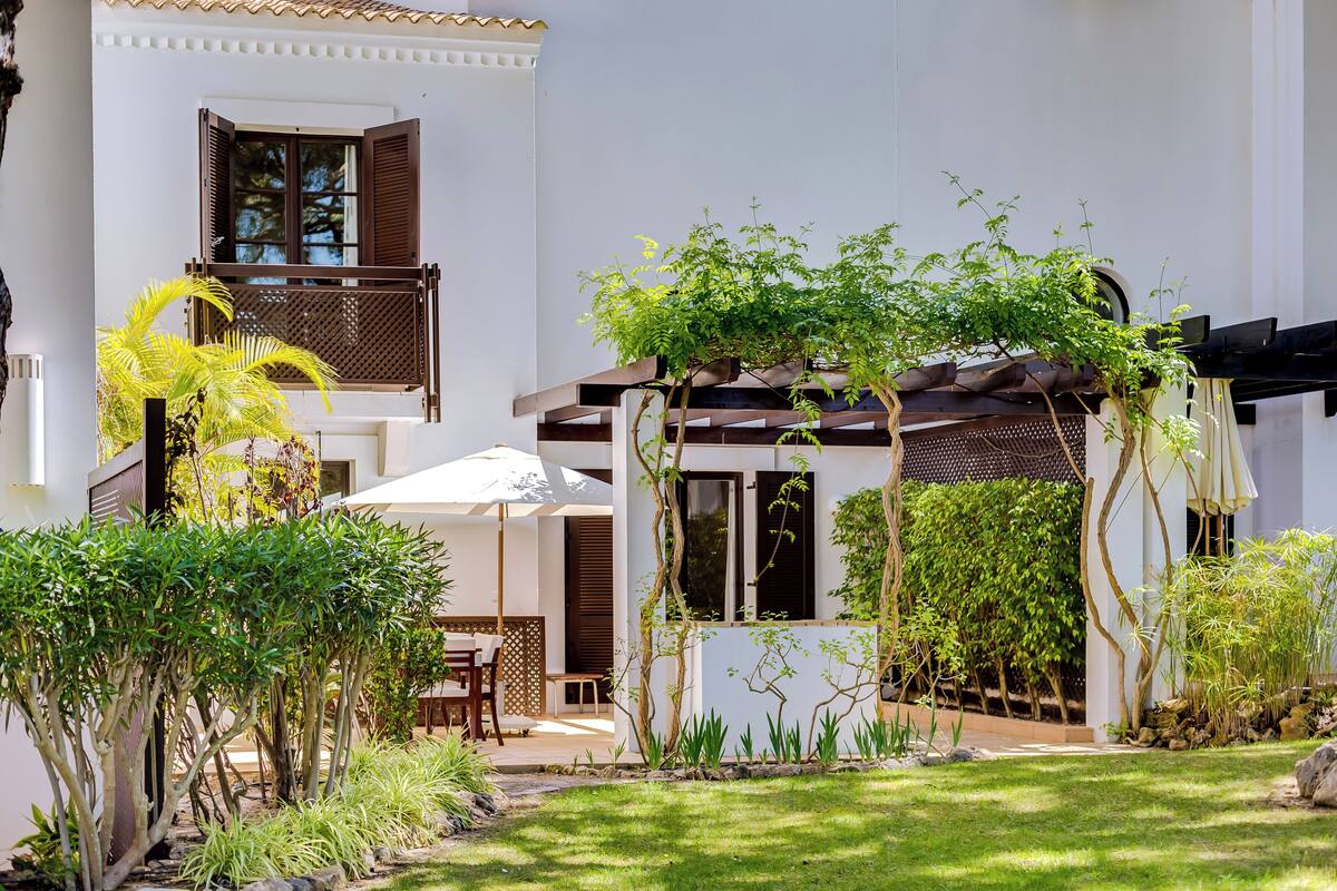 A white building with a balcony, a covered outdoor seating area, and various plants.
