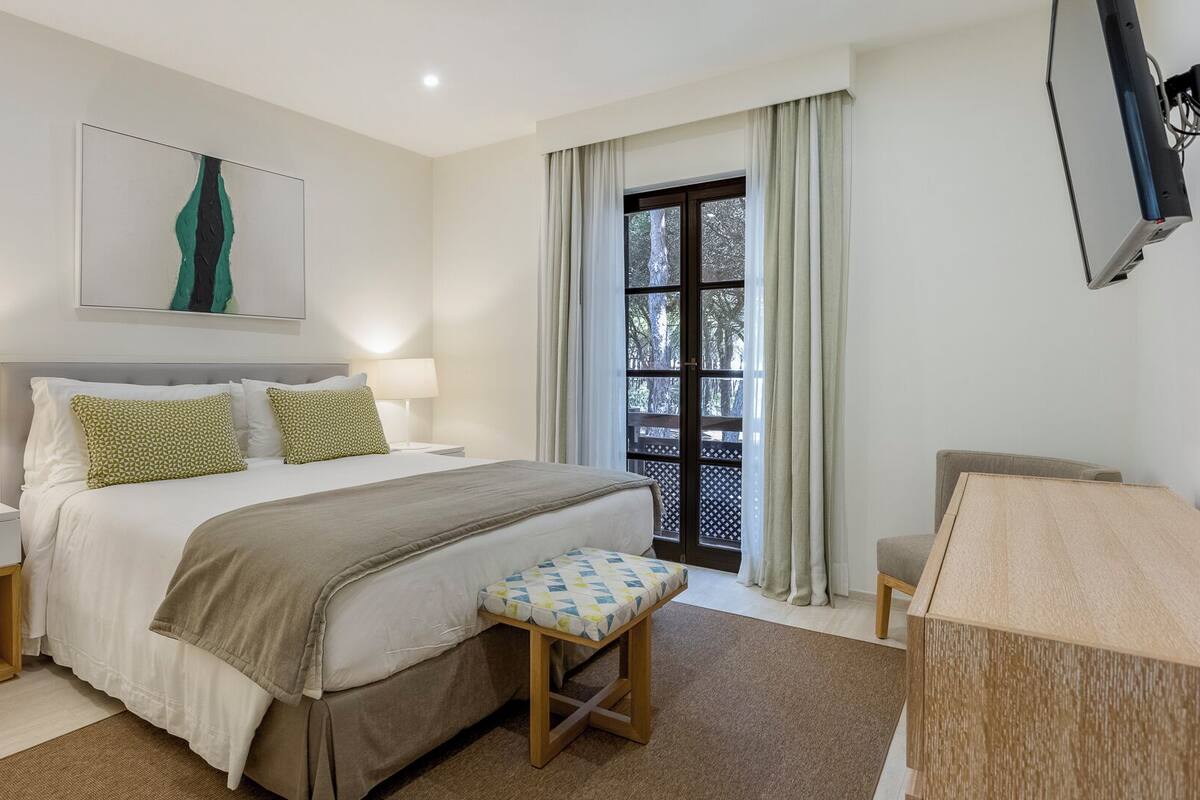 A modern hotel bedroom with a large bed, bedside tables, a bench, a TV, and a window with curtains.