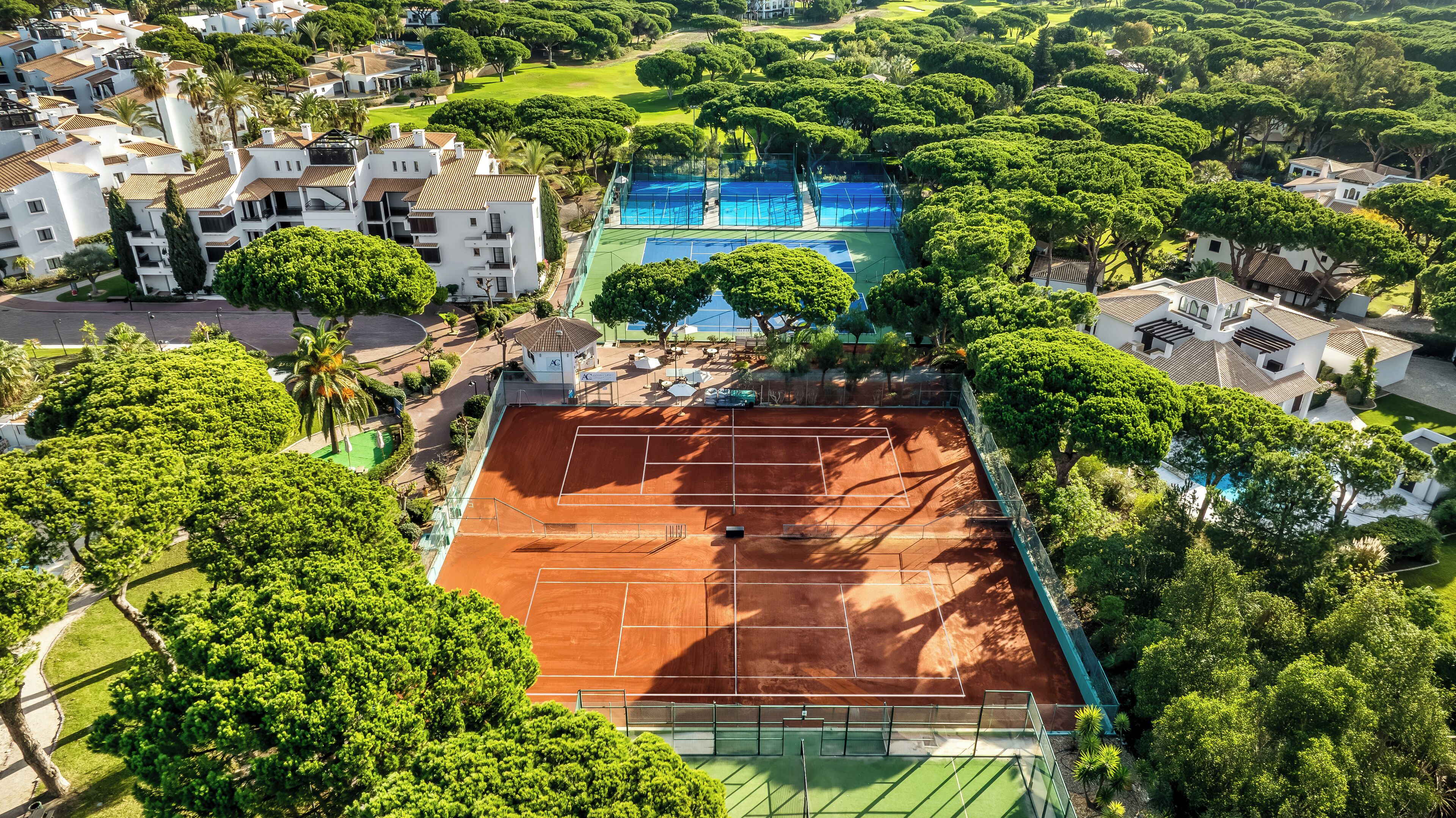 An aerial view of a residential area with a tennis court surrounded by lush greenery and white buildings with red-tiled roofs.